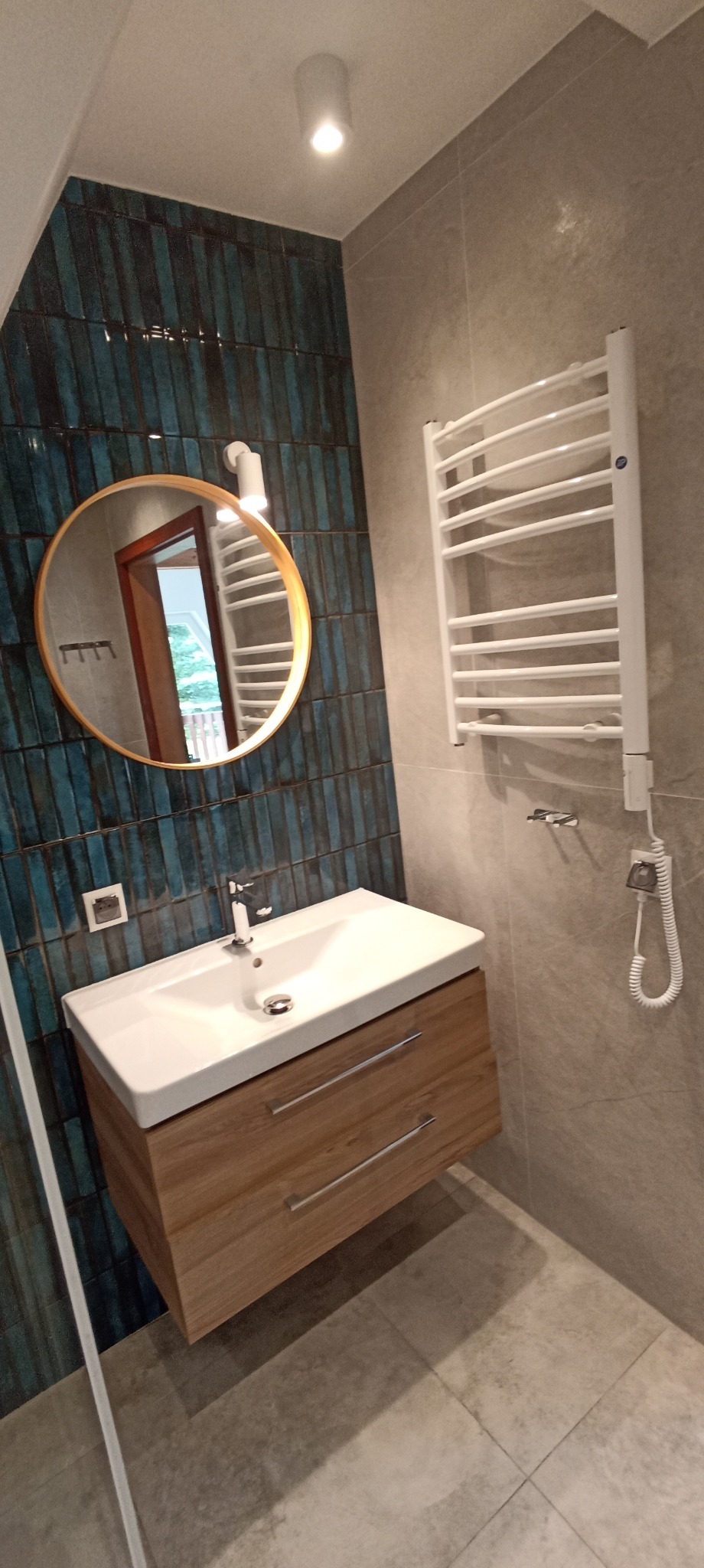 Nowoczesna łazienka: umywalka z drewnianą szafką, okrągłe lustro z oświetleniem, niebieska glazura i biały grzejnik drabinkowy na szarej ścianie.