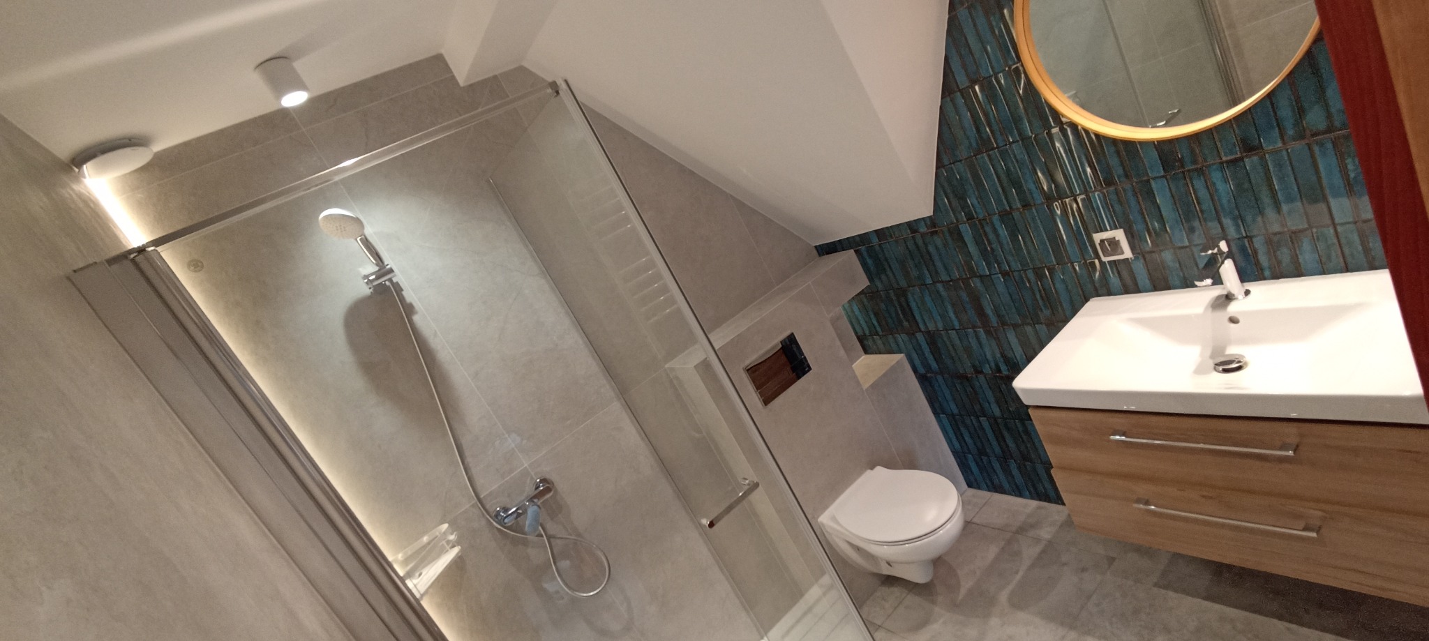 Nowoczesna łazienka na poddaszu z kabiną prysznicową, umywalką z szafką z drewna i toaletą. Ściana zdobiona turkusowymi płytkami w pionowy wzór.