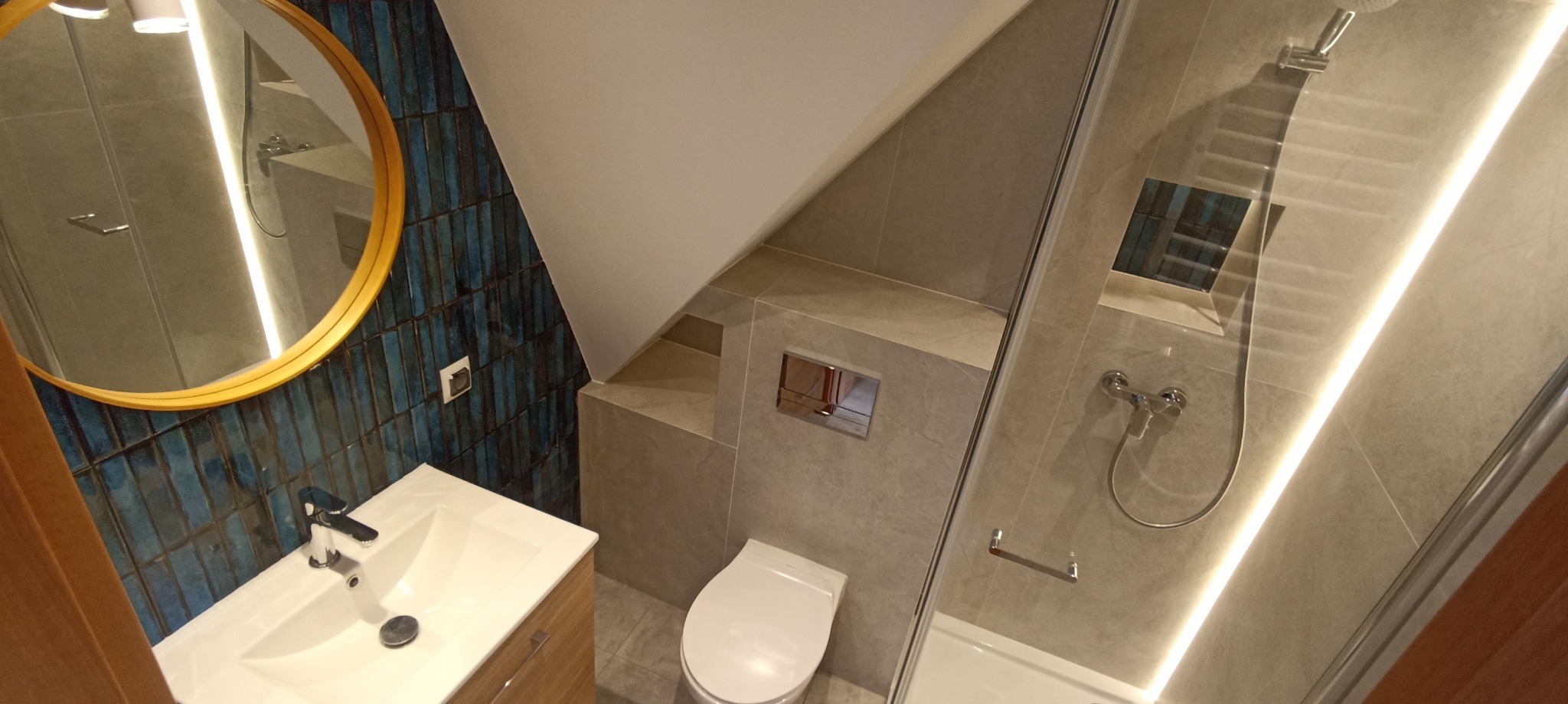 Nowoczesna łazienka na poddaszu:  niebieska ściana z lustrem, umywalka z szafką, toaleta i kabina prysznicowa z podświetleniem LED.