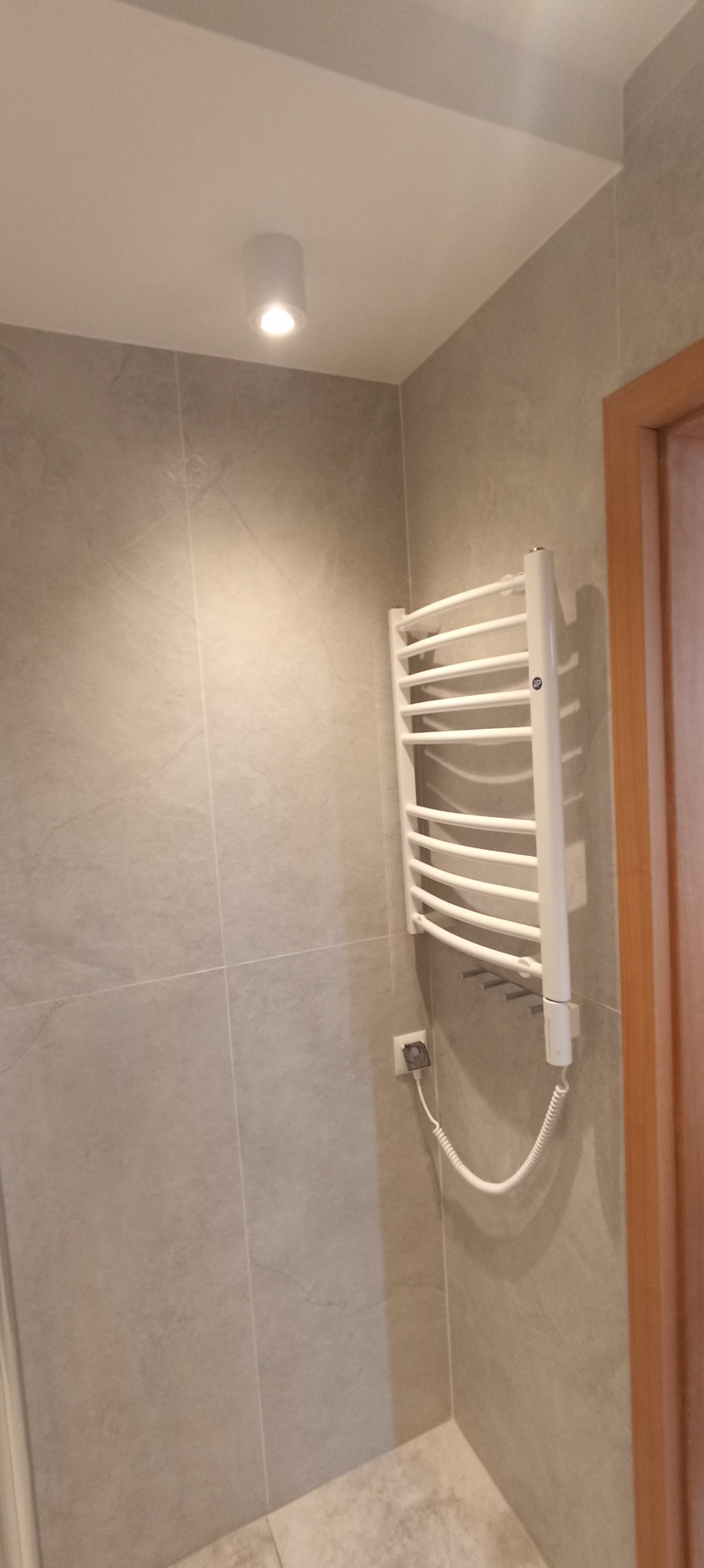 Wykończona łazienka z grzejnikiem drabinkowym, szare płytki na ścianach i podłodze, biała sufitowa lampa punktowa, fragment drewnianej ościeżnicy drzwi.