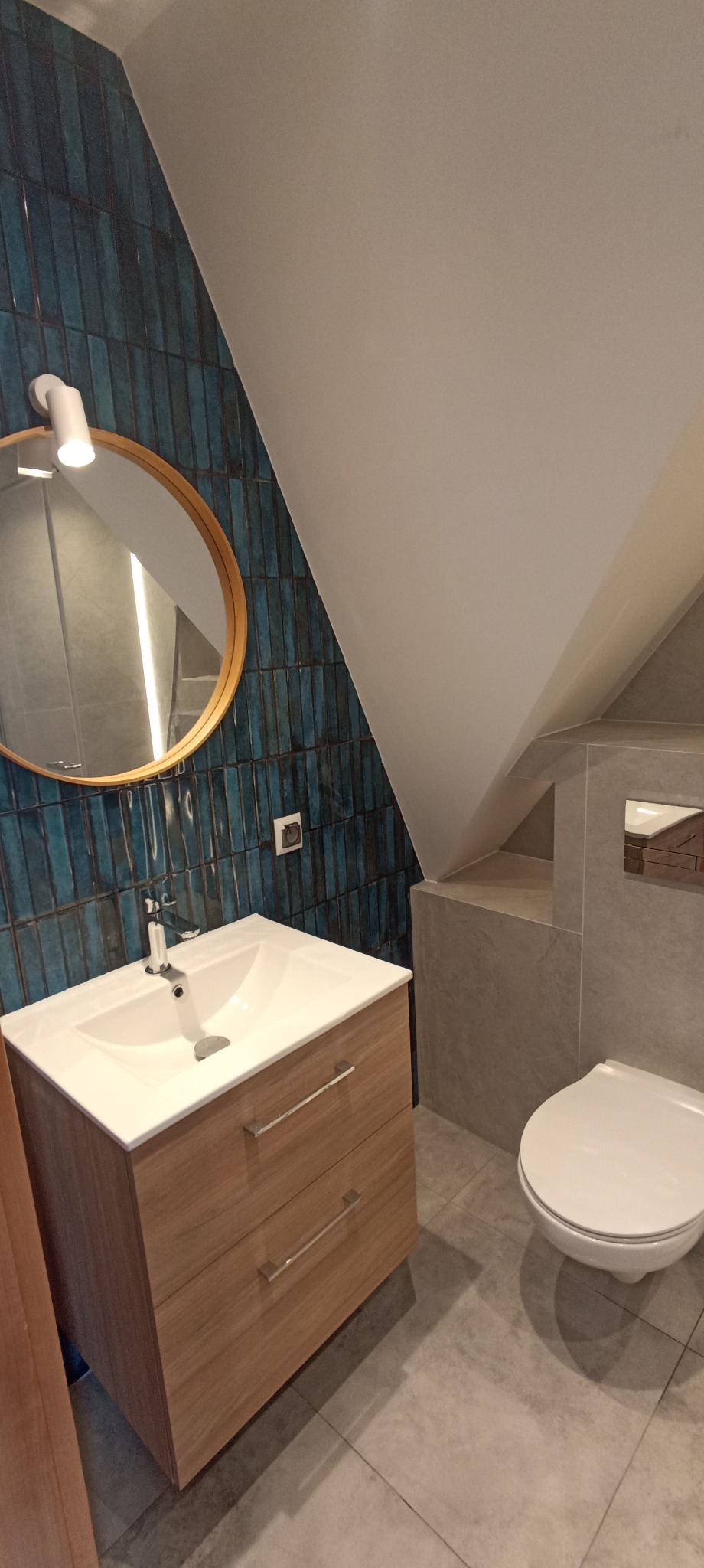 Łazienka na poddaszu z umywalką w szafce z drewna, toaletą w szarości i ścianą z niebieskich płytek. Okrągłe lustro z podświetleniem.