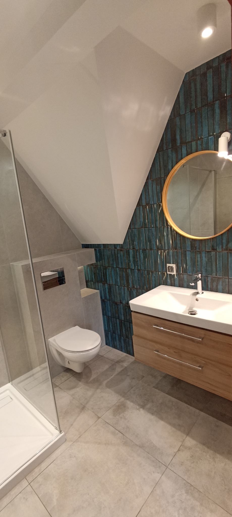 Nowoczesna łazienka na poddaszu z kabiną prysznicową, toaletą podwieszaną i umywalką z szafką. Ściana wyłożona pionowymi, niebieskimi płytkami.