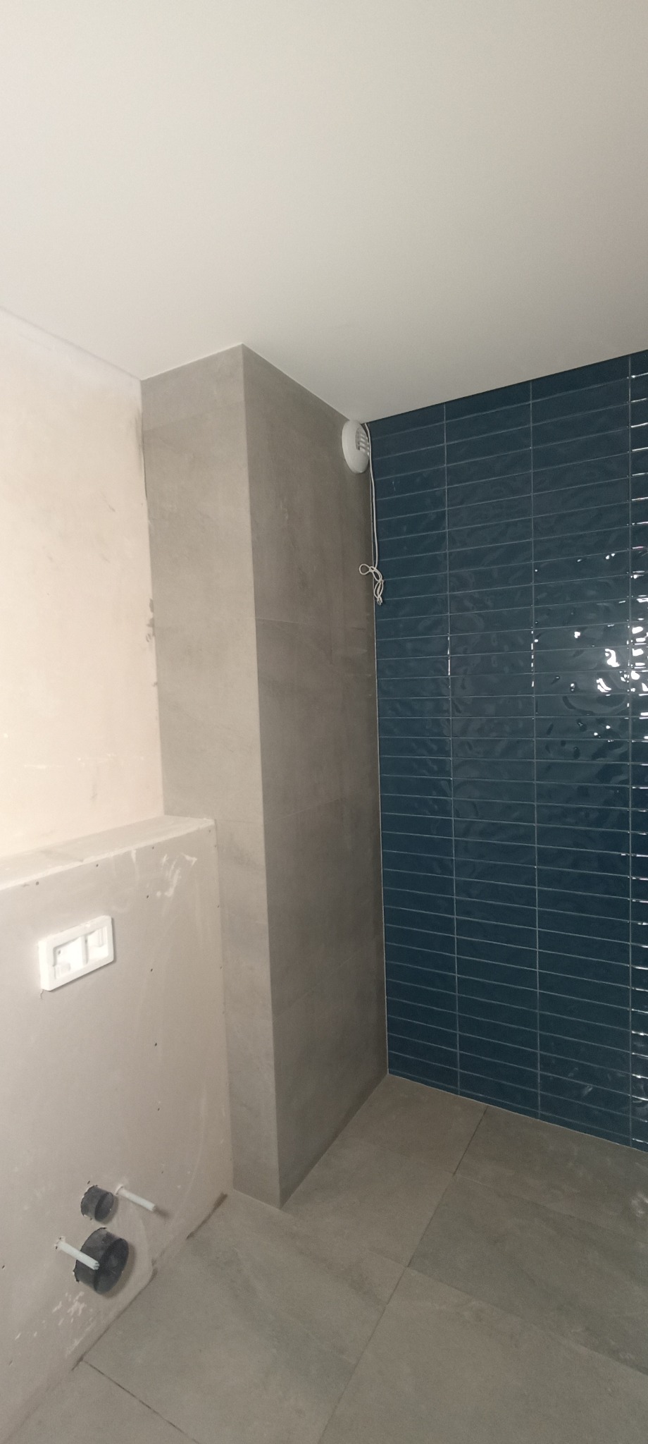 Wykończona ściana z granatowej, błyszczącej glazury w kształcie cegiełek, szara kolumna z płytek oraz fragment ściany przed wykończeniem w łazience.