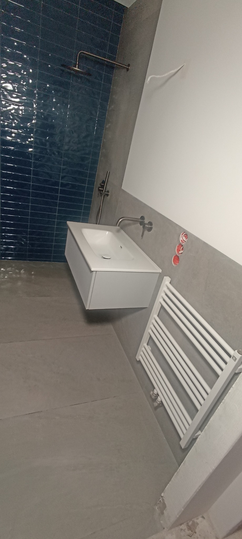 Nowoczesna łazienka po remoncie: ciemnoniebieskie płytki na ścianie pod prysznicem, biała umywalka z szafką i grzejnik drabinkowy. Minimalistyczny design.