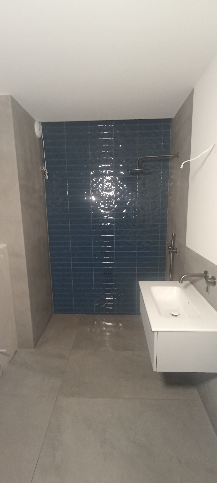 Wykończona łazienka z umywalką w szafce, kabiną prysznicową z ciemnoniebieską glazurą i szarą podłogą z płytek. Widoczne baterie i elementy instalacji.