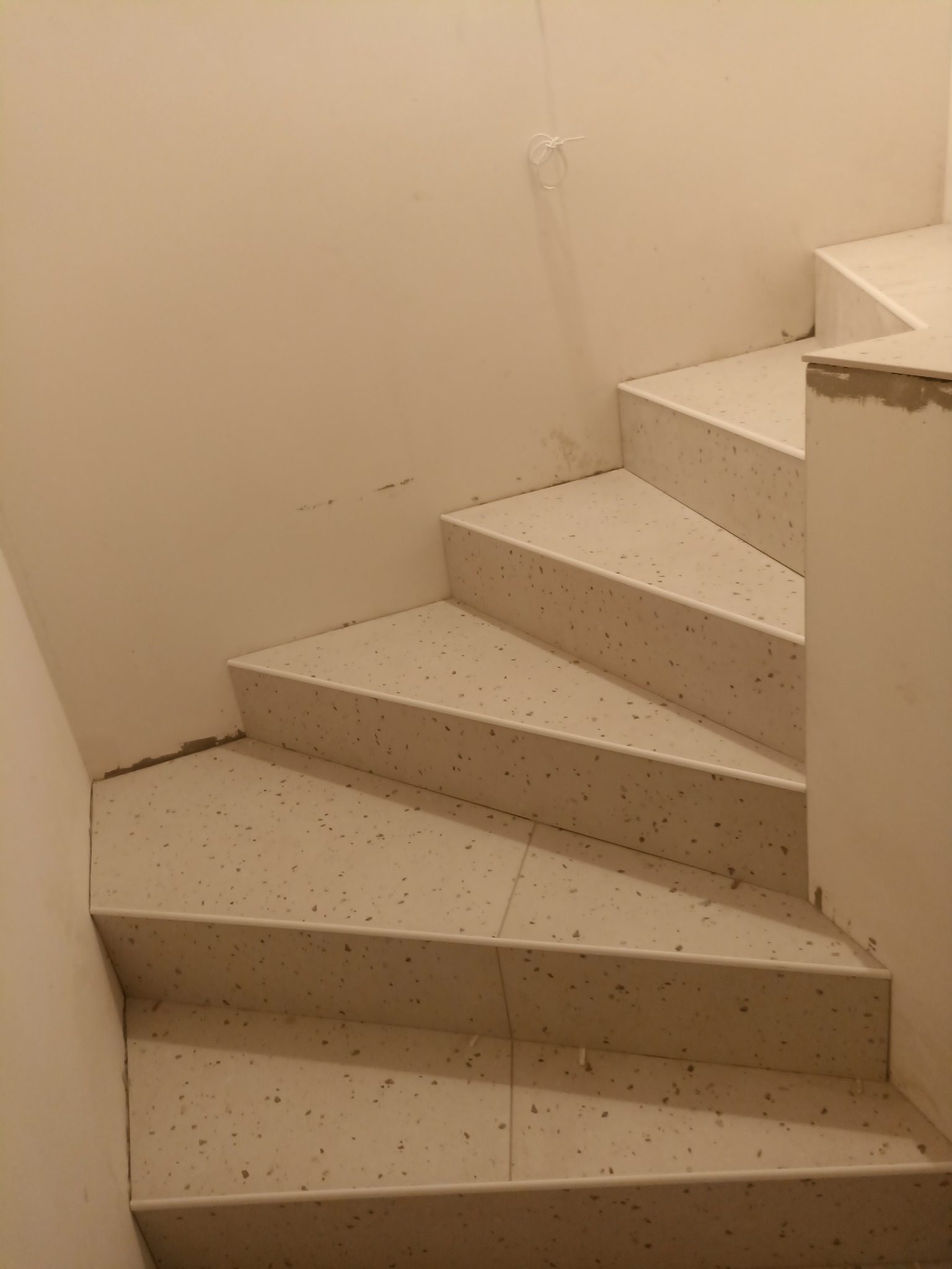 Wykończone schody wewnętrzne z jasnych płytek lastryko, widok z góry. Ściana w kolorze złamanej bieli z widocznym fragmentem instalacji elektrycznej.