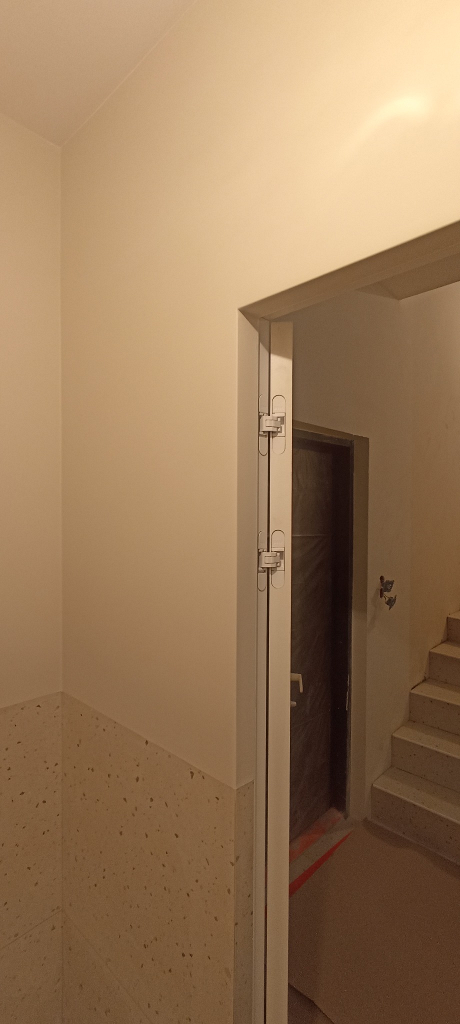 Wykończone wnętrze z widoczną ścianą w jasnym odcieniu, fragmentem schodów i drzwiami z ukrytymi zawiasami. Wykończenie z terazzo na dolnej części ściany.