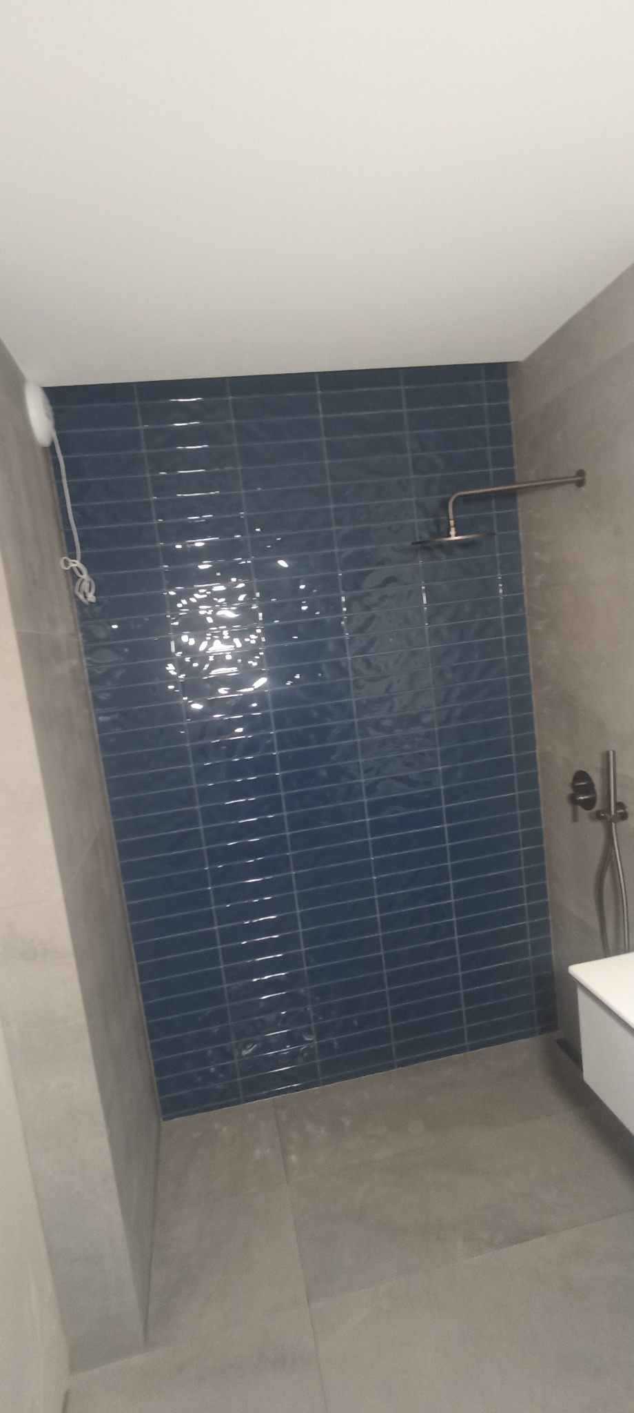 Niebieskie, błyszczące płytki w kabinie prysznicowej, szare ściany i podłoga. Widoczna bateria i deszczownica. Minimalistyczny design łazienki.