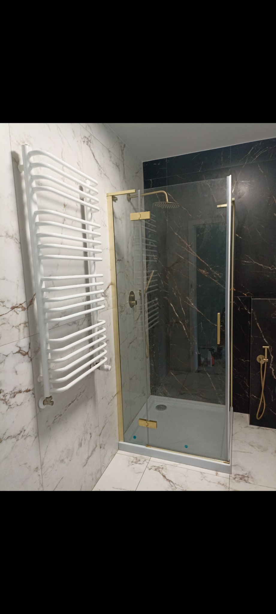Nowoczesna kabina prysznicowa z złotymi detalami i biały grzejnik na tle marmurowych płytek w wykończonej łazience.