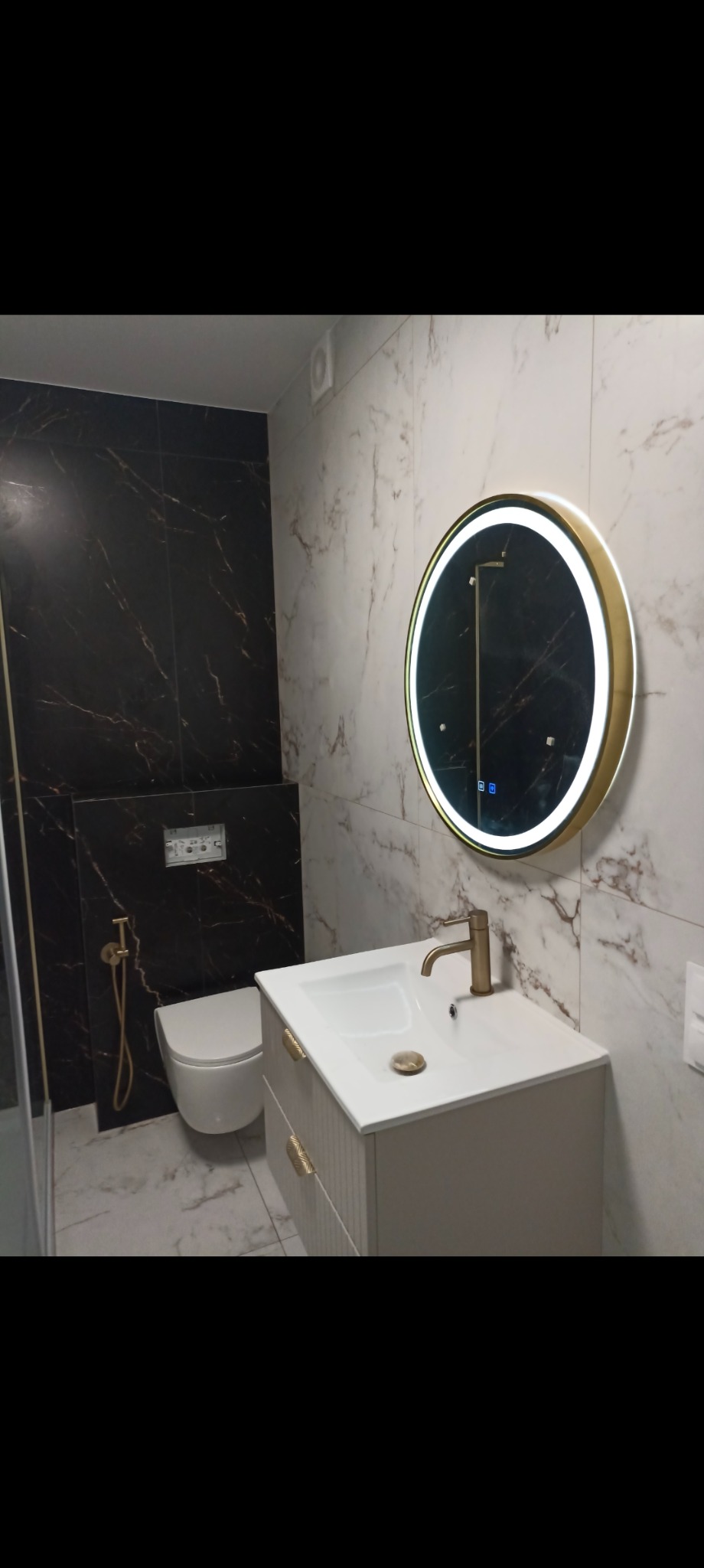 Elegancka łazienka po remoncie: biała szafka z umywalką, okrągłe lustro z podświetleniem LED, toaleta podwieszana i czarno-białe marmurowe płytki.