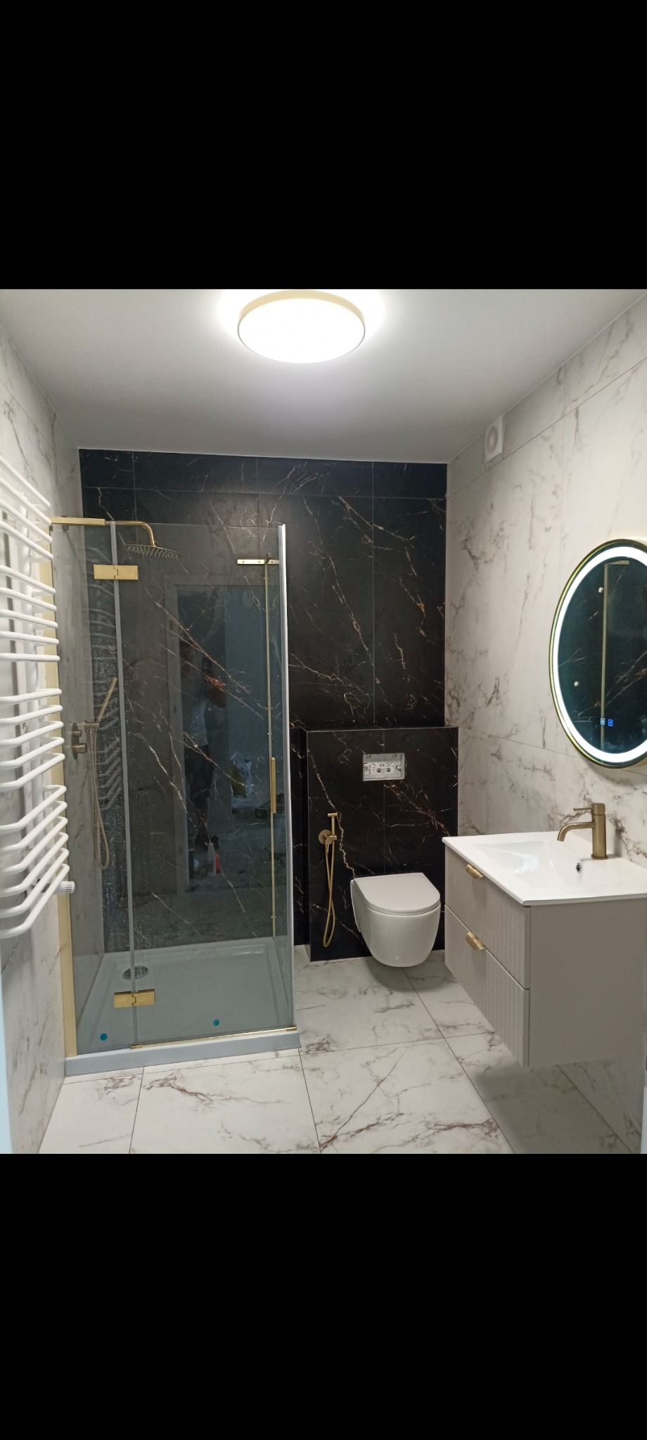 Elegancka łazienka z kabiną prysznicową typu walk-in, umywalką z szafką i podświetlanym lustrem. Czarne i białe marmurowe płytki tworzą kontrastowe wykończenie.