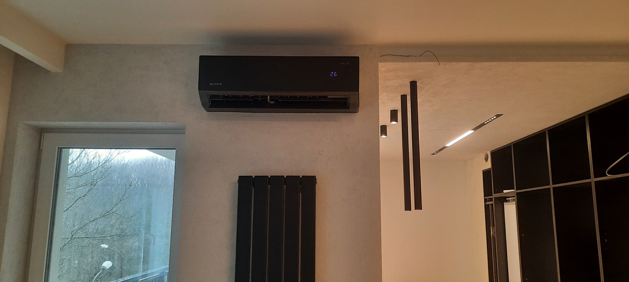 Czarny klimatyzator ścienny marki Rotenso w nowoczesnym wnętrzu z dekoracyjną ścianą, obok okna i czarnego grzejnika.