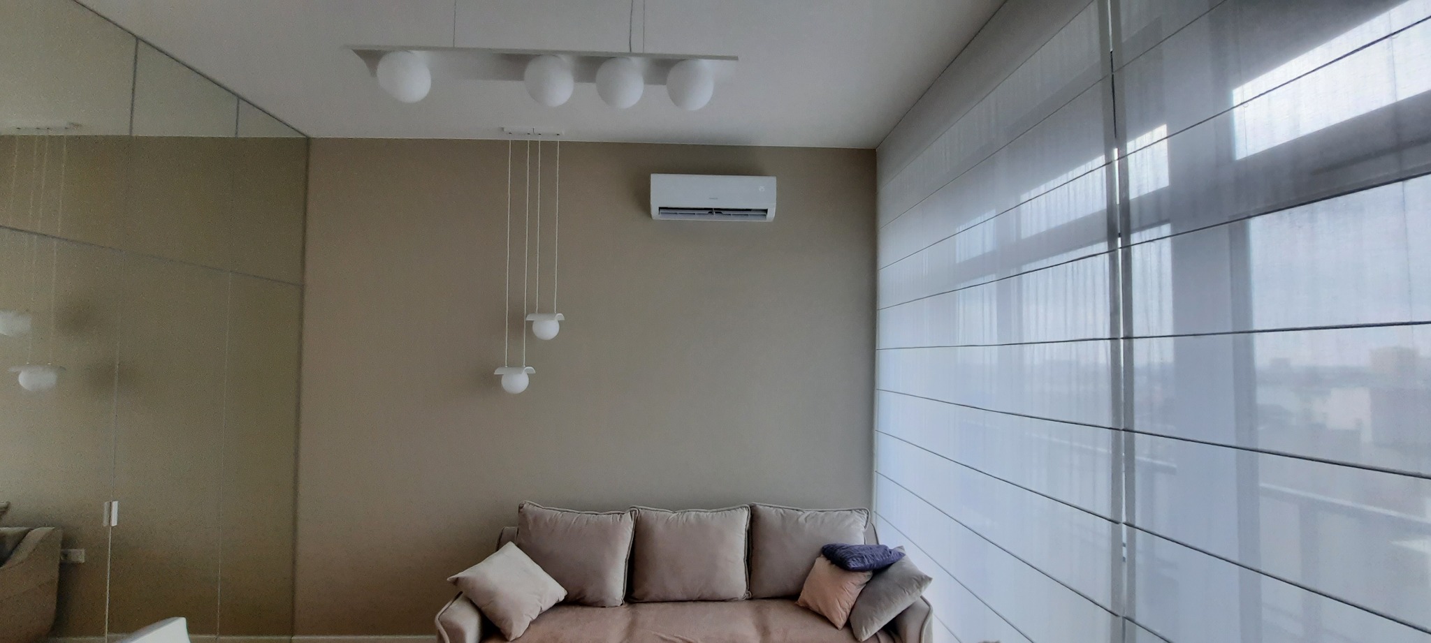 Jasne wnętrze salonu z beżową sofą, zasłoniętym oknem i zamontowaną klimatyzacją pod sufitem, oświetlone wiszącymi lampami.
