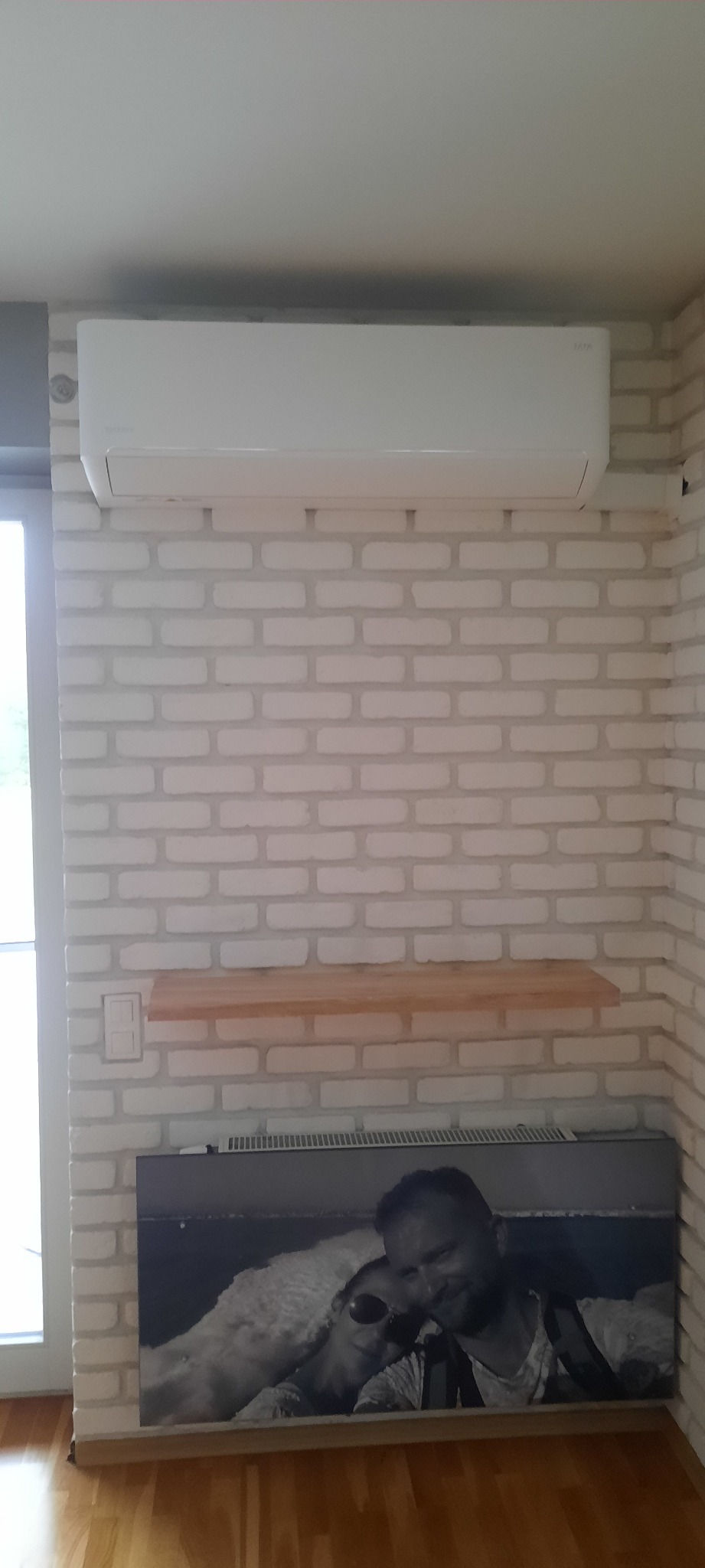 Biała klimatyzacja zamontowana na ścianie z białej cegły, pod nią półka i obraz z ludźmi.