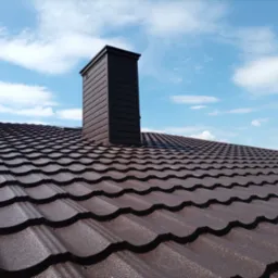 Brązowy dach z blachy trapezowej z kominem na tle błękitnego nieba z chmurami.