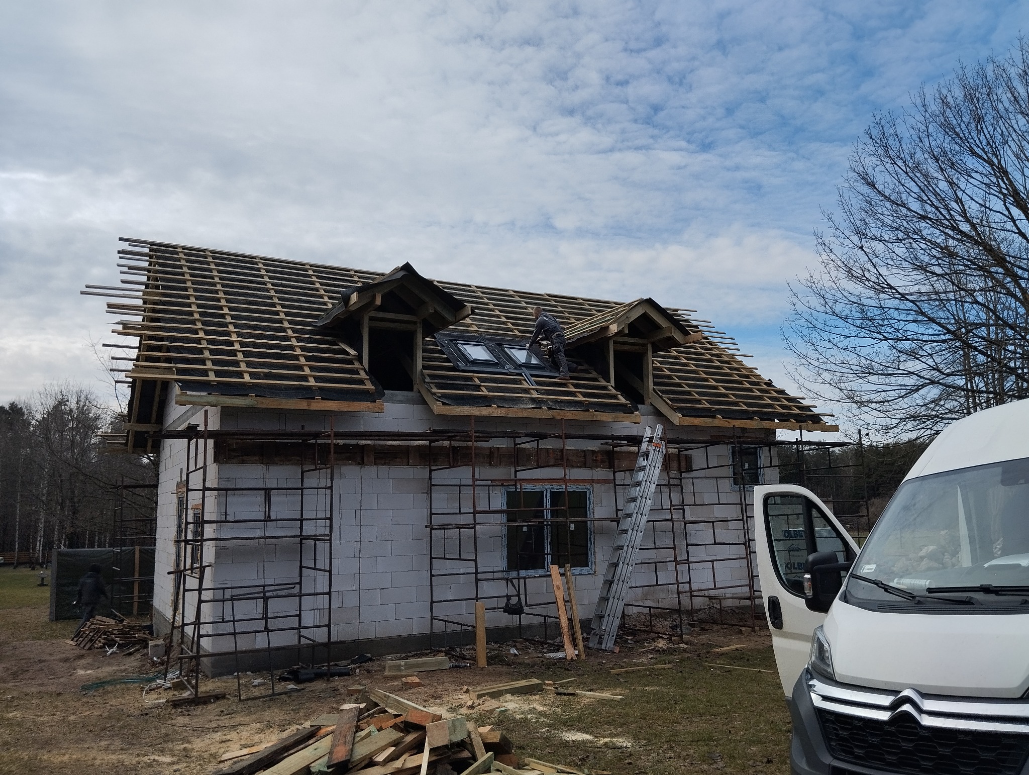 Budowa domu jednorodzinnego w stanie surowym otwartym: widoczna więźba dachowa z oknami dachowymi montowanymi przez pracownika, rusztowania i materiały budowlane na posesji.