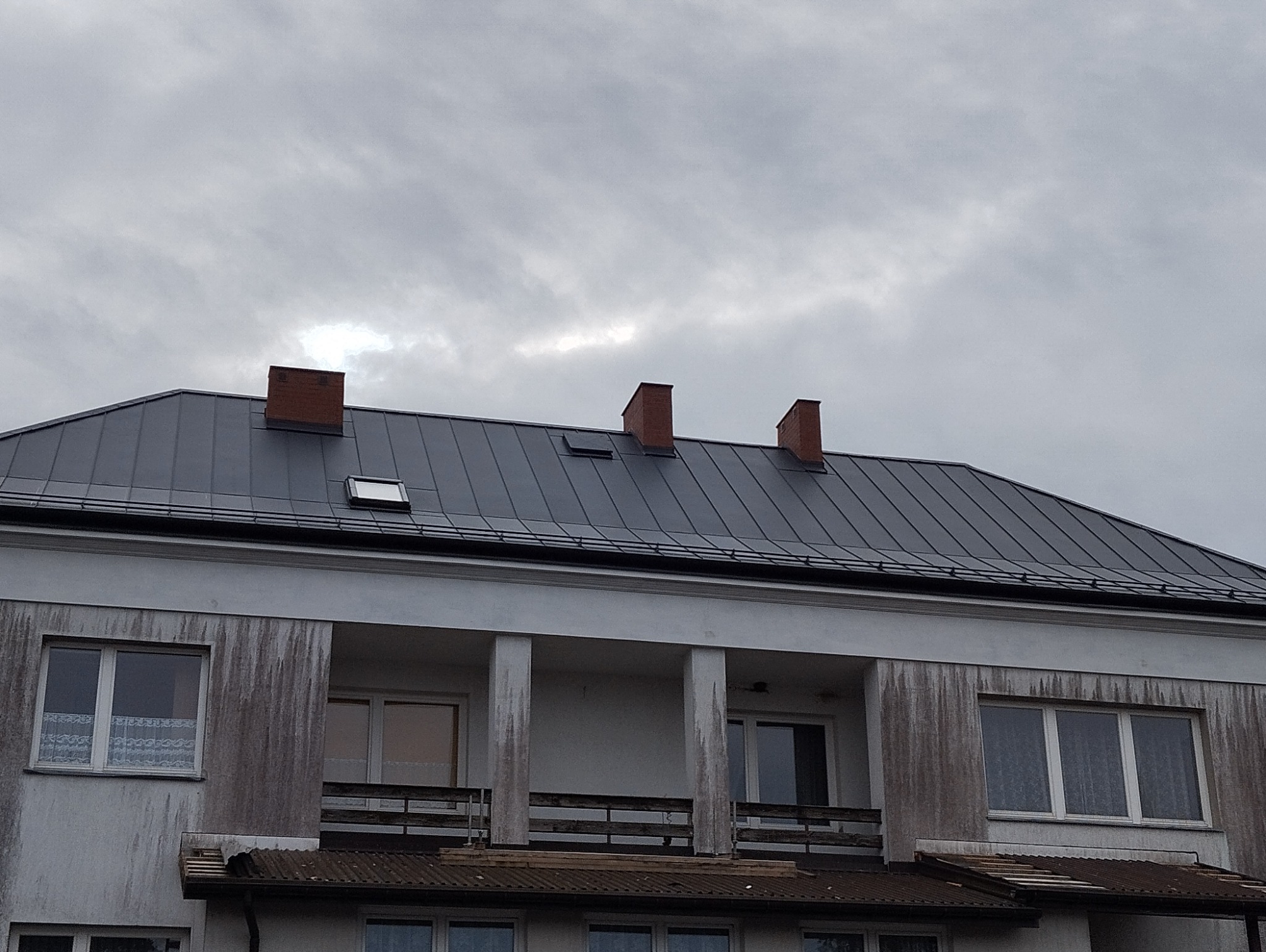 Szary, blaszany dach budynku z trzema kominami z cegły i oknem dachowym, na tle pochmurnego nieba.