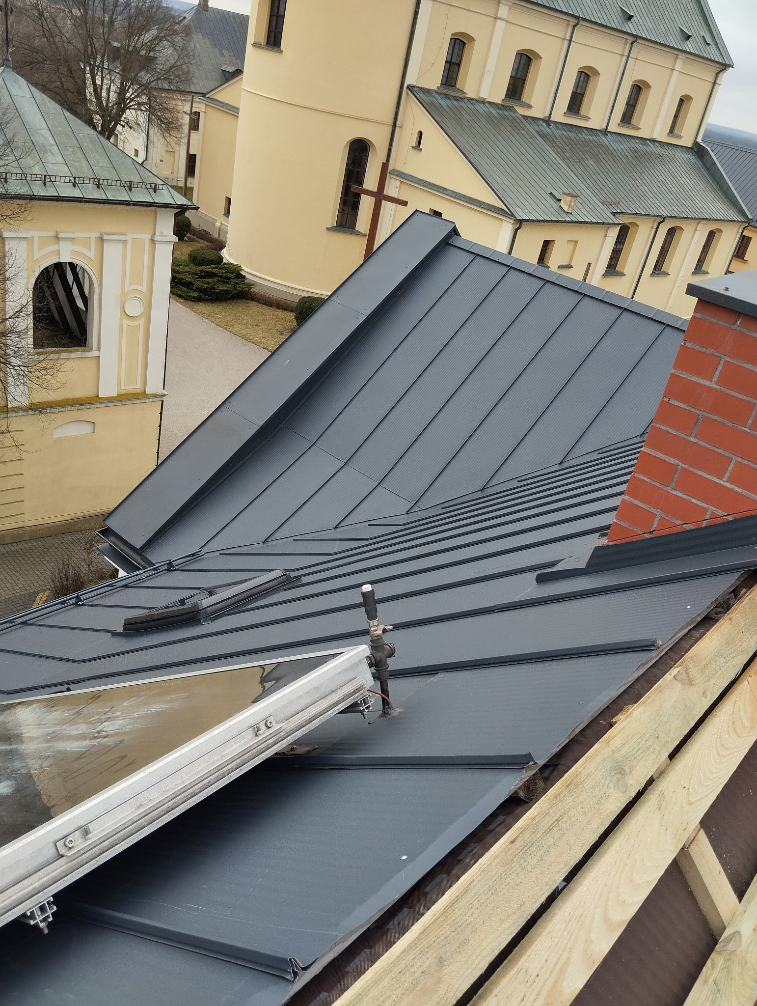 Szare blaszane pokrycie dachu z oknem dachowym w trakcie montażu, widoczny komin z czerwonej cegły oraz elementy więźby dachowej, w tle budynek kościoła.