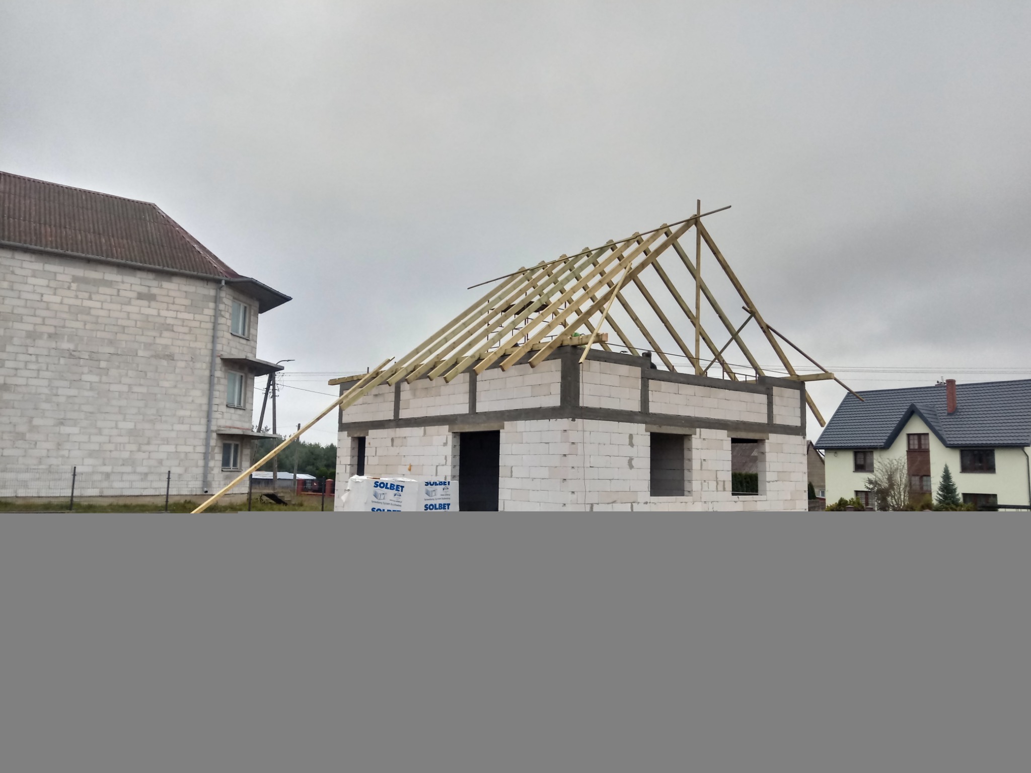 Surowa konstrukcja domu z białych pustaków z częściowo wykonaną drewnianą konstrukcją dachu na tle pochmurnego nieba i sąsiednich budynków.
