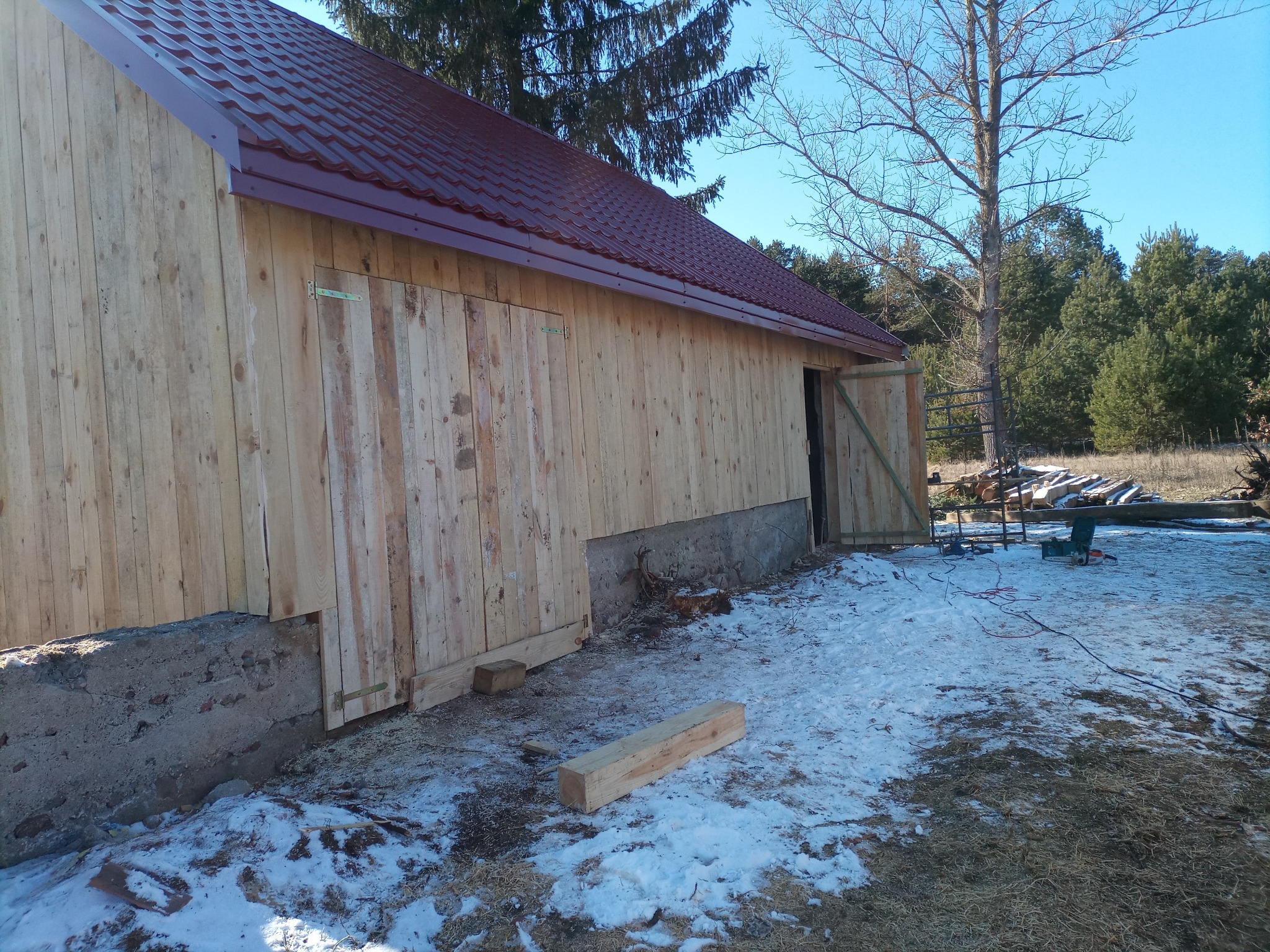 Budowa drewnianego budynku gospodarczego z czerwonym dachem w Siemiatyczach, widoczne deski elewacyjne, fundament i częściowo pokryty dach, na ziemi leży śnieg i drewniane elementy.