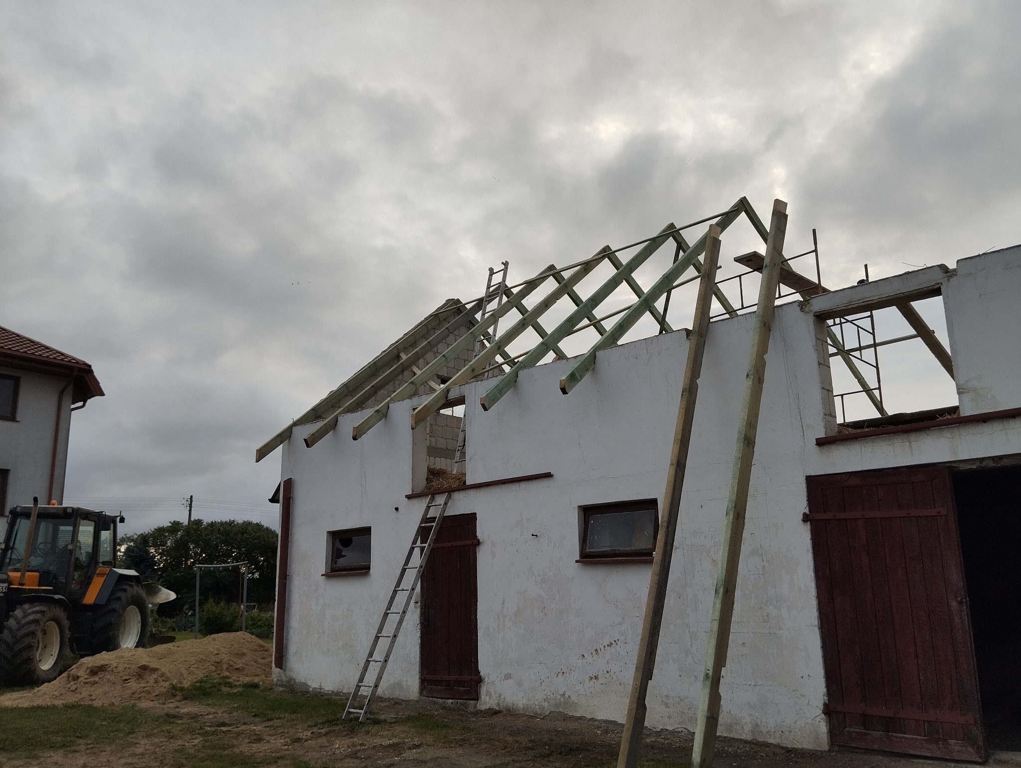 Budynek gospodarczy w trakcie budowy dachu z drewnianą więźbą, widoczna drabina oparta o ścianę, traktor w tle i pochmurne niebo.