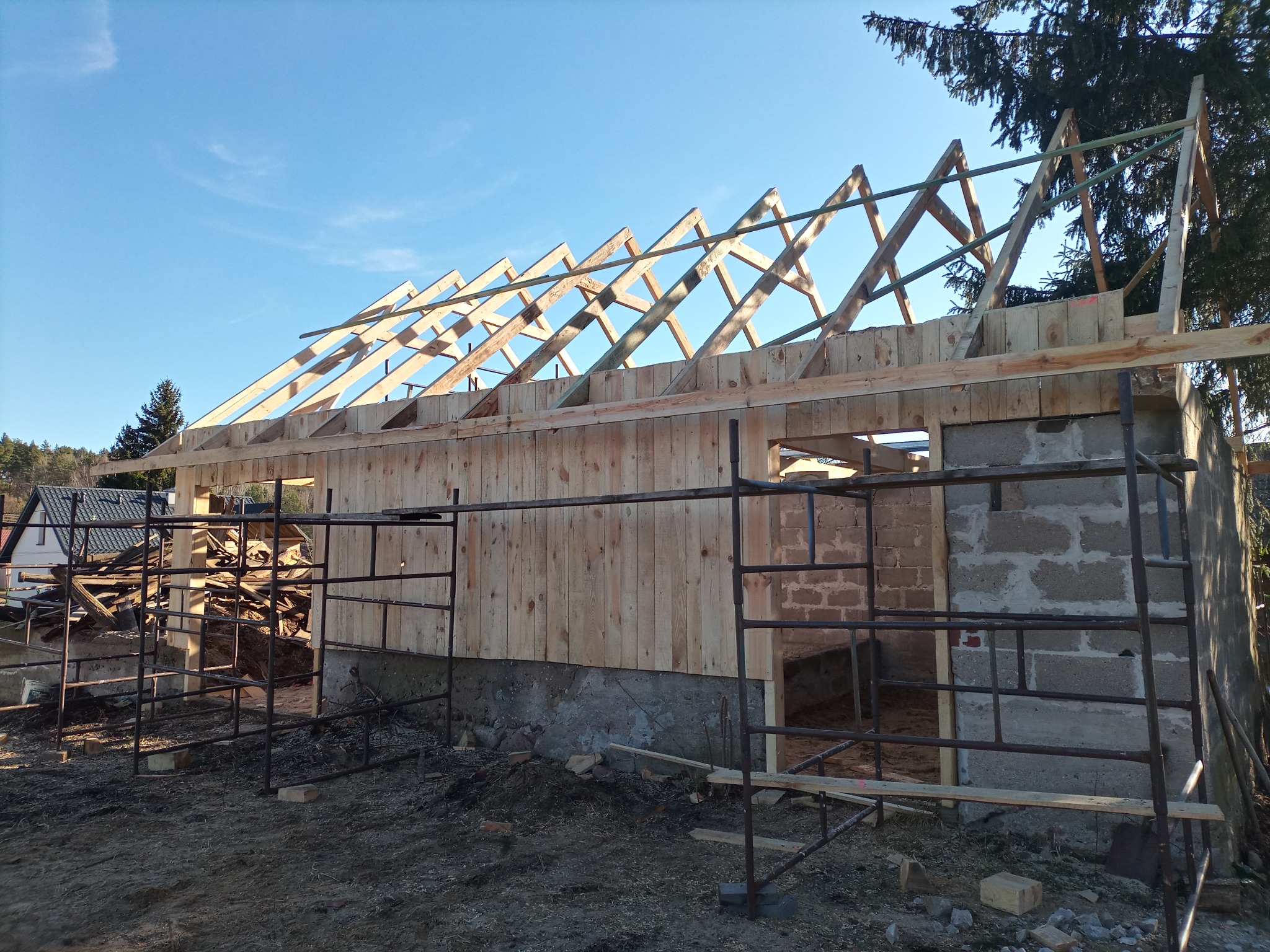 Wznoszenie drewnianej więźby dachowej na murowanym budynku gospodarczym, widoczne rusztowania i niebieskie niebo.