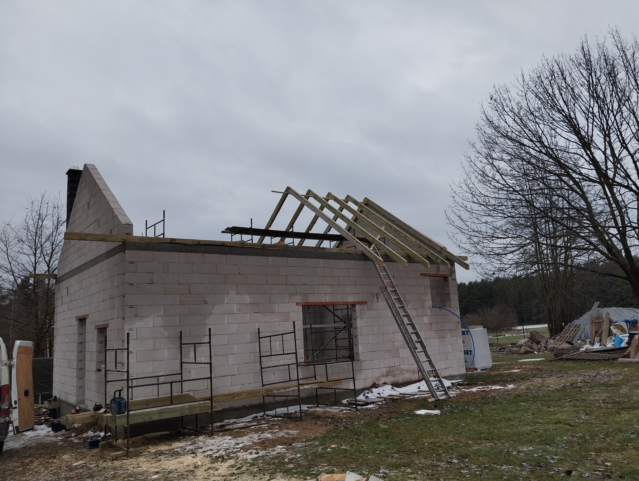 Budowa domu z widoczną konstrukcją więźby dachowej, ściany z białych bloczków, rusztowania i drabina oparta o budynek w pochmurny dzień.