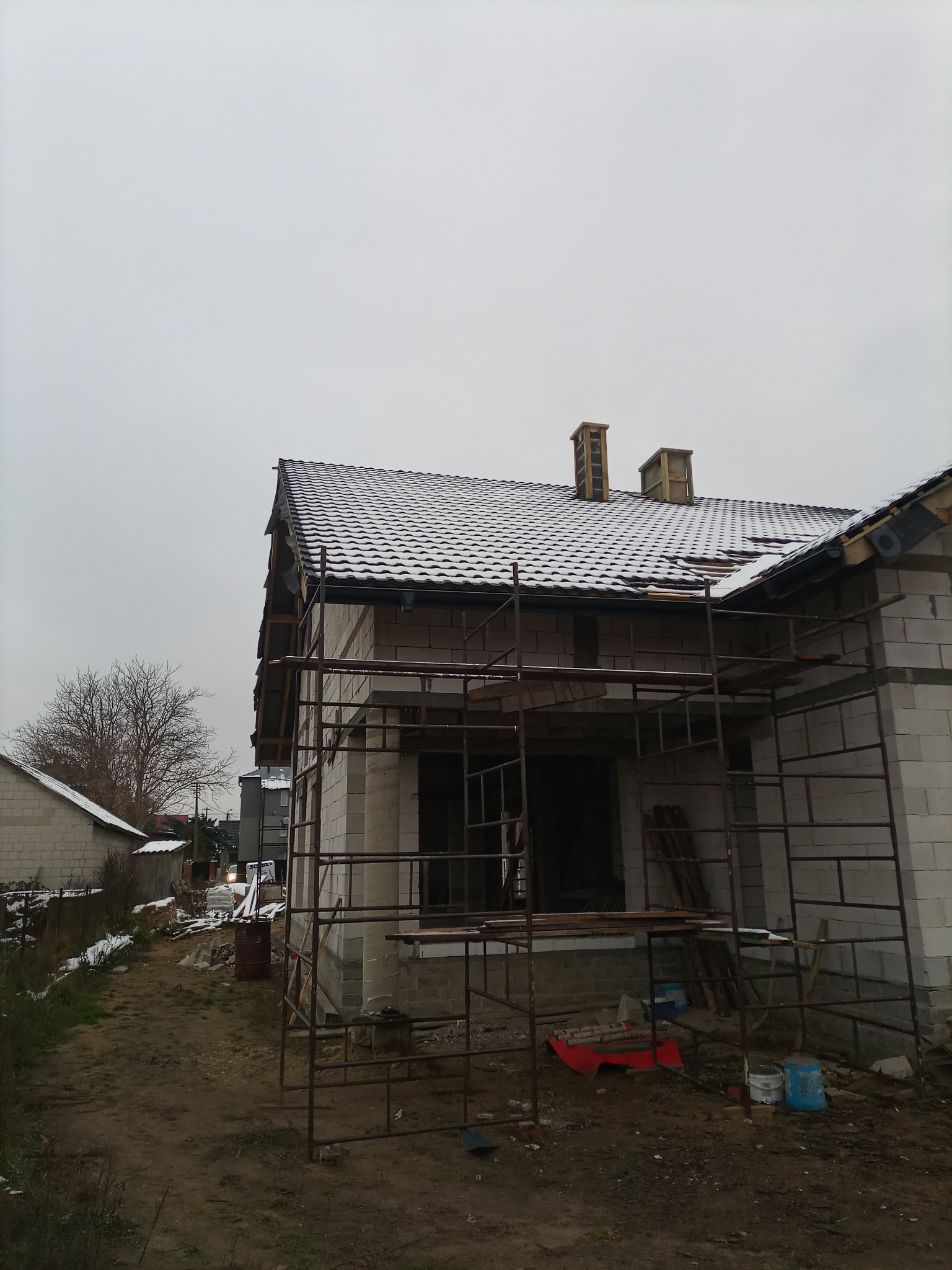 Dom w trakcie budowy z częściowo położonym dachem pokrytym śniegiem, widoczne rusztowania i materiały budowlane.