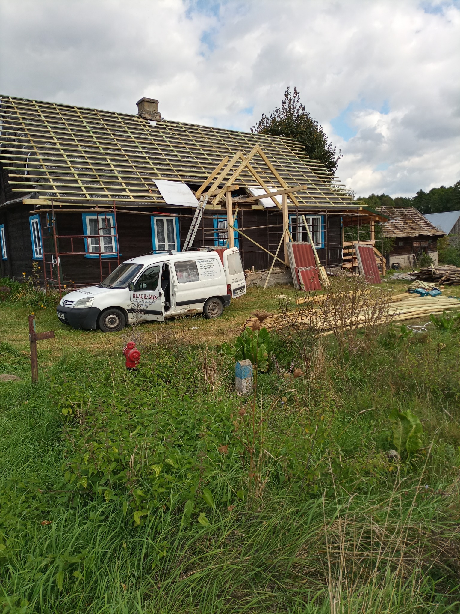 Drewniany dom w trakcie remontu dachu, widoczna więźba dachowa, częściowo pokryta czarną membraną, na podwórku samochód dostawczy firmy dekarskiej, materiały budowlane i rusztowania.