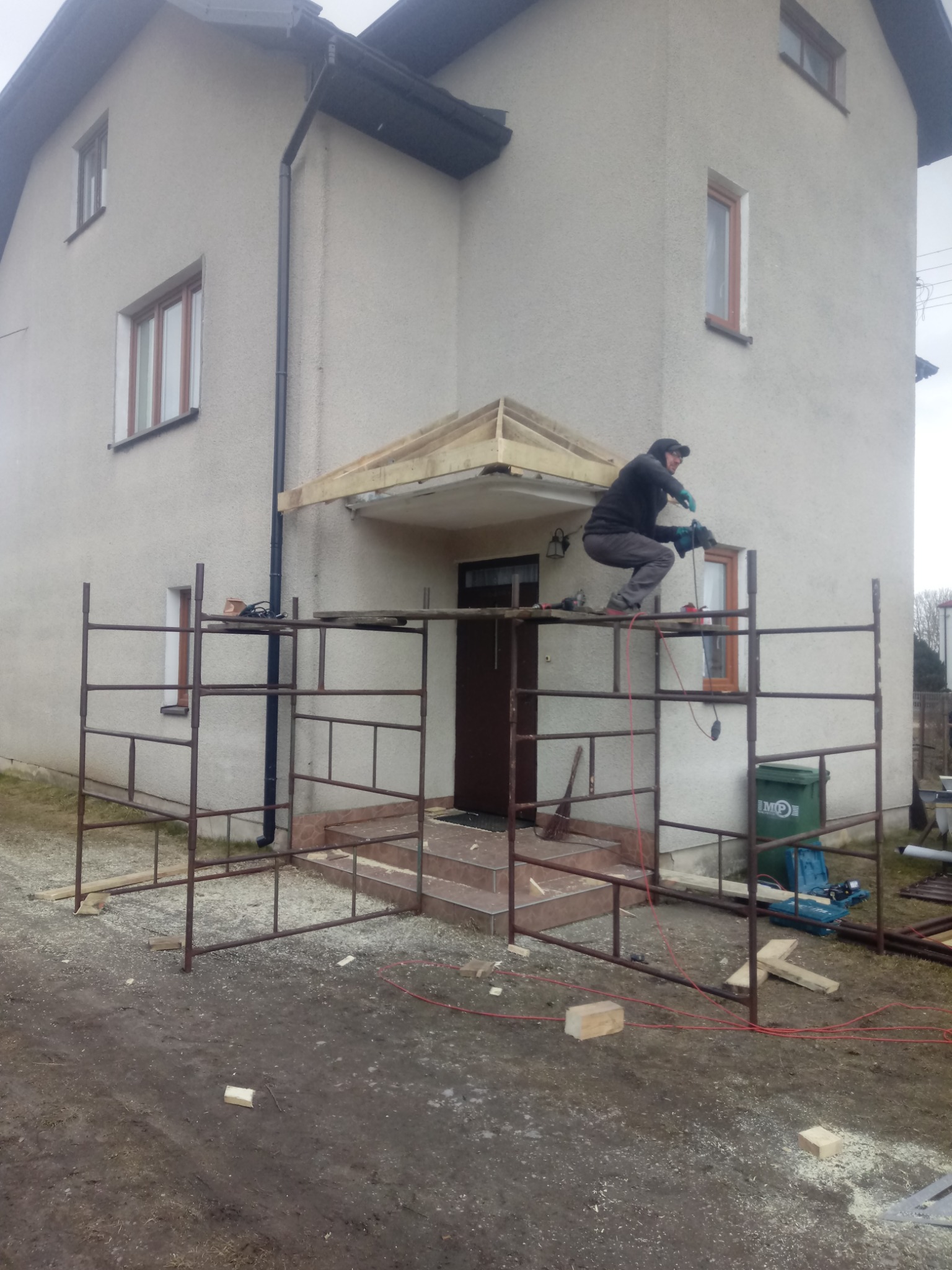 Pracownik na rusztowaniu, w trakcie montażu drewnianego zadaszenia nad wejściem do domu, używa wiertarki; widoczne narzędzia i elementy konstrukcyjne.