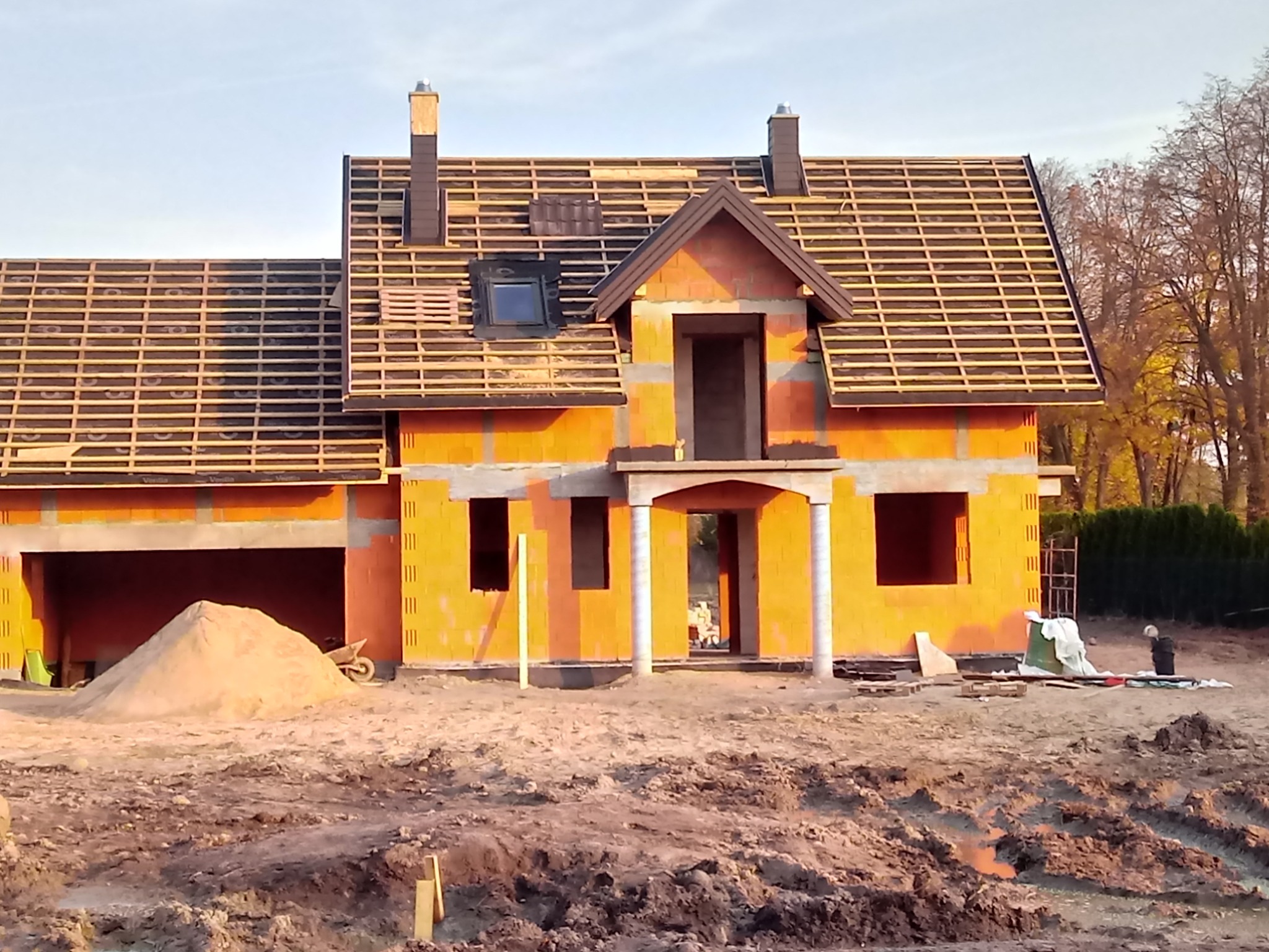 Dom w trakcie budowy z widoczną konstrukcją dachu, częściowo pokrytą łatami i kontrłatami; widoczne okno dachowe i komin, na pierwszym planie hałda piasku i teren budowy.