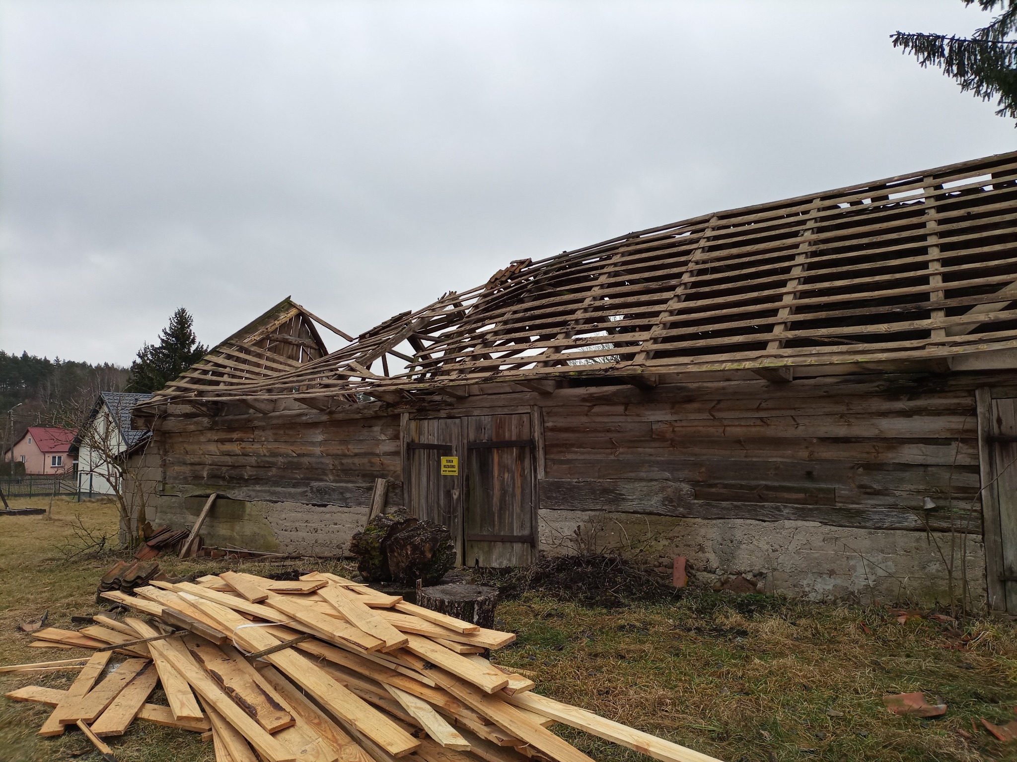 Stary drewniany budynek gospodarczy z częściowo rozebraną konstrukcją dachu, widoczne deski i elementy więźby, na pierwszym planie stos desek, pochmurne niebo w tle.