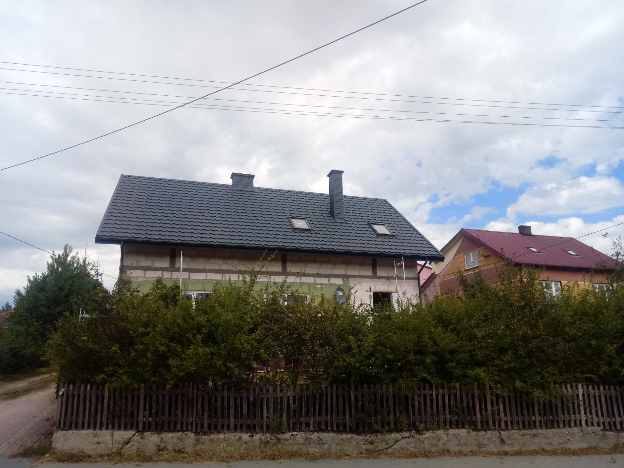 Dom jednorodzinny w trakcie budowy z nowym, ciemnym dachem z dachówki modułowej, widoczne kominy i okna dachowe, elewacja w surowym stanie, częściowo zasłonięta krzewami i drewnianym płotem.
