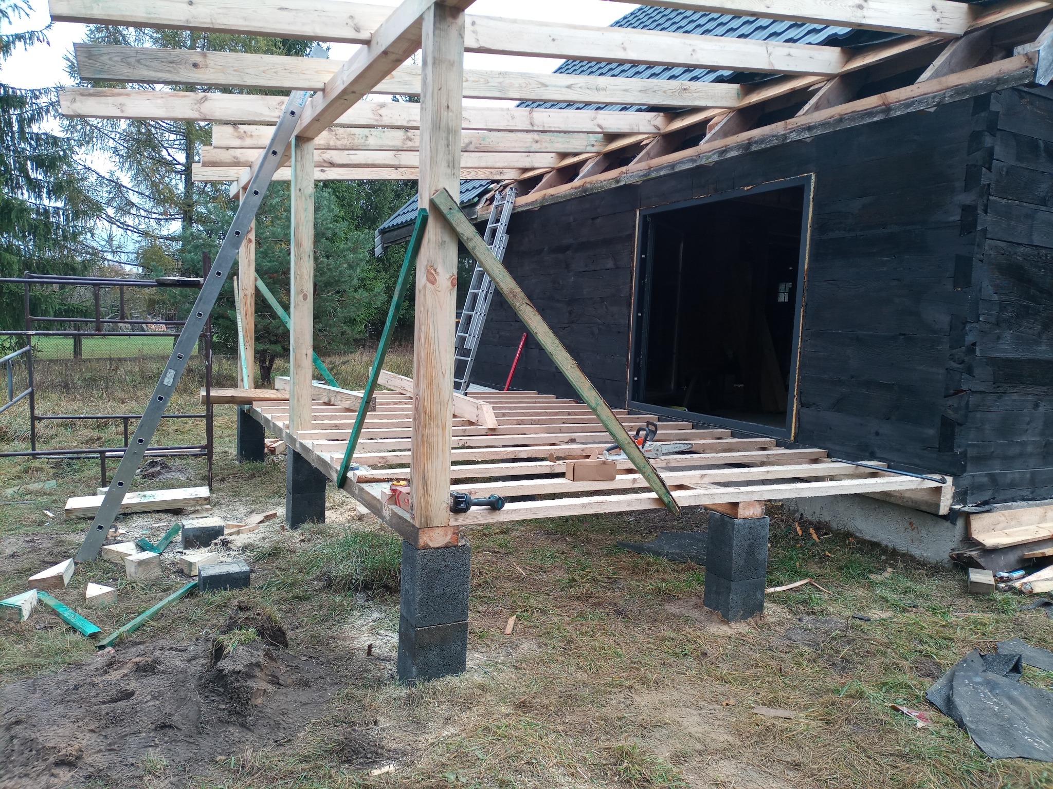 Wznoszenie drewnianej konstrukcji dachu na czarnej, drewnianej chacie, widoczne elementy więźby dachowej, drabina oparta o konstrukcję, teren budowy z resztkami materiałów.