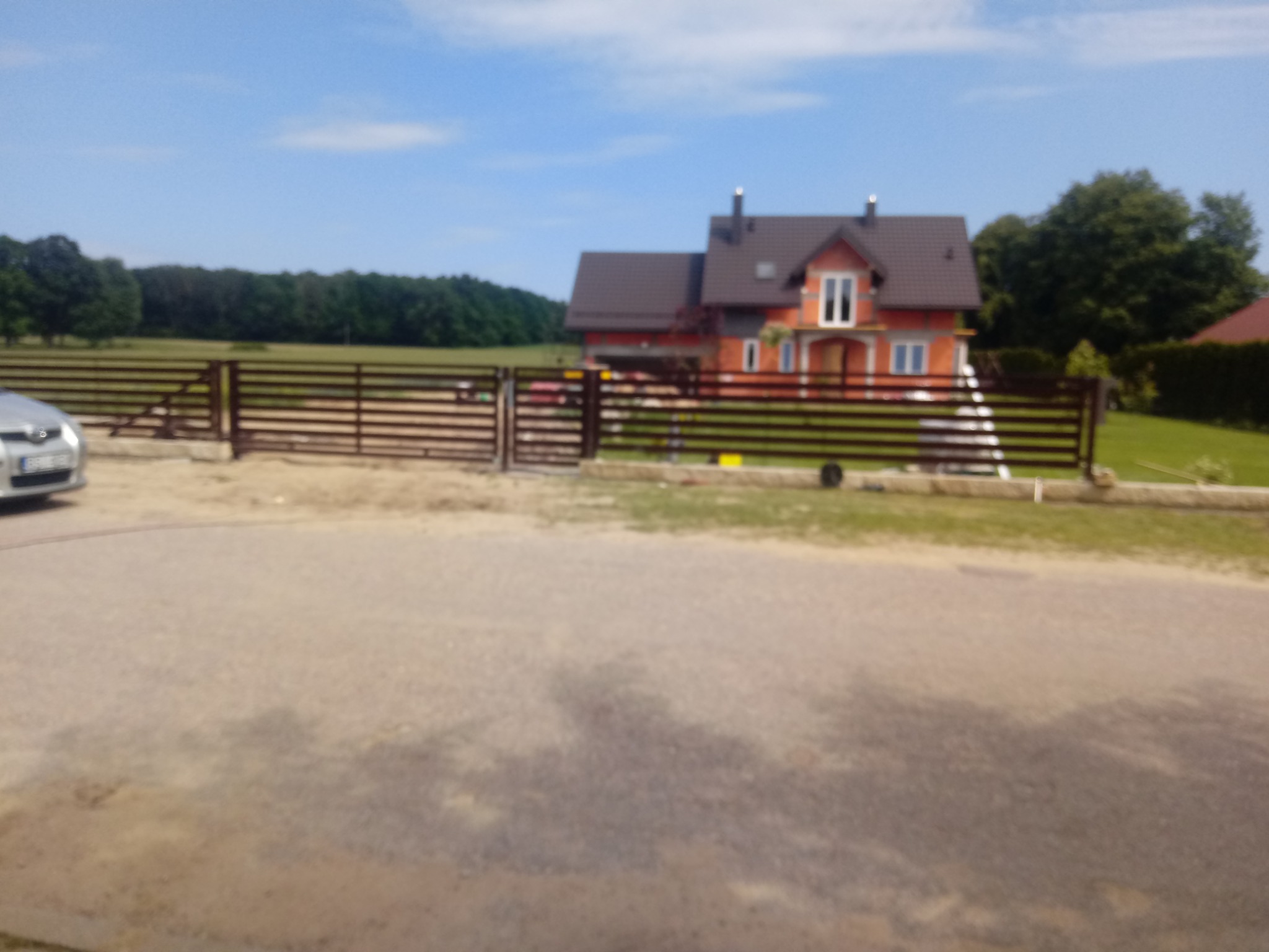 Dom jednorodzinny z ciemnobrązowym dachem i jasnymi oknami, widziany zza drewnianego ogrodzenia, z samochodem w tle po lewej stronie.