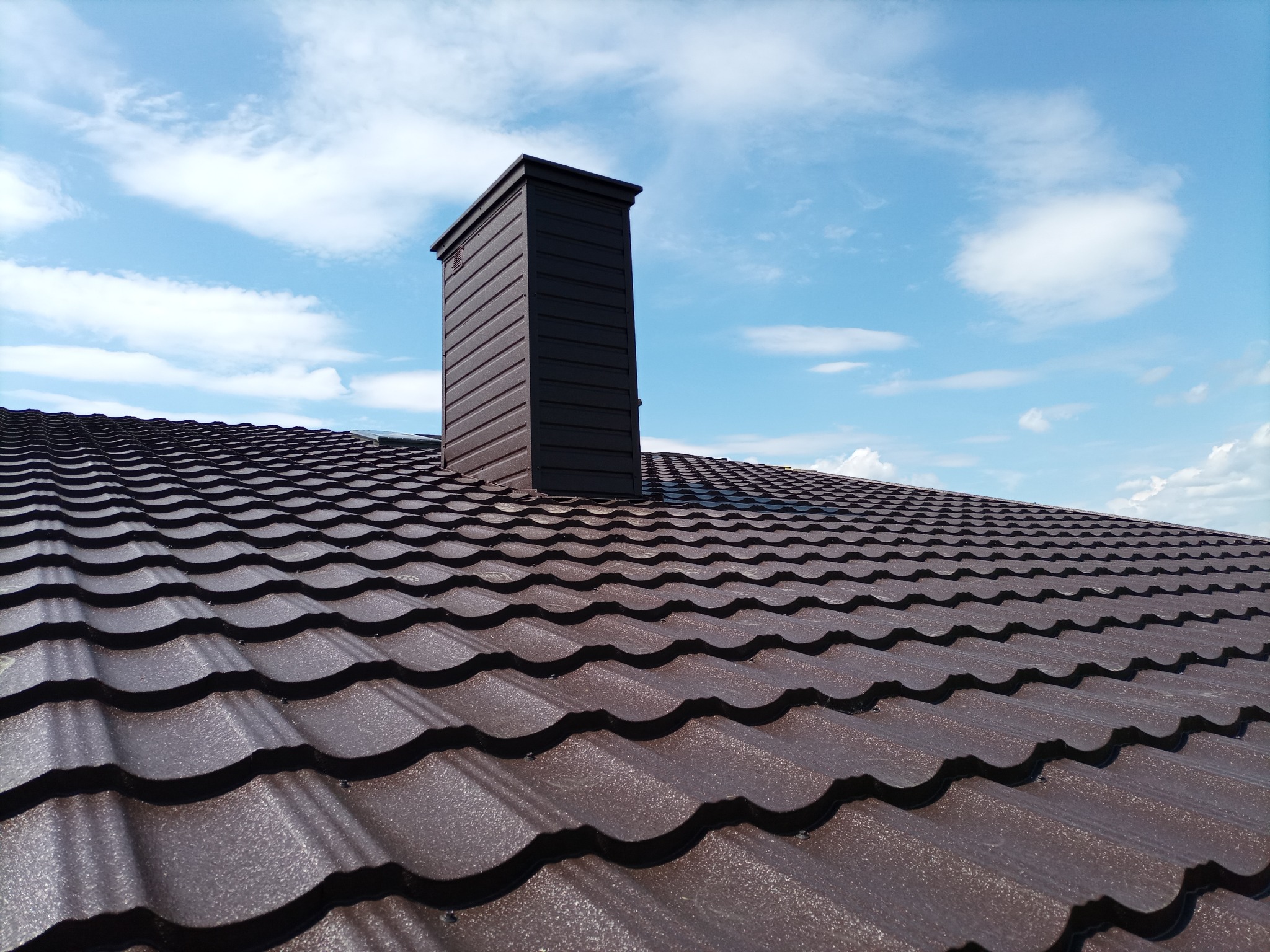 Brązowy dach z blachy trapezowej z kominem na tle błękitnego nieba z chmurami.