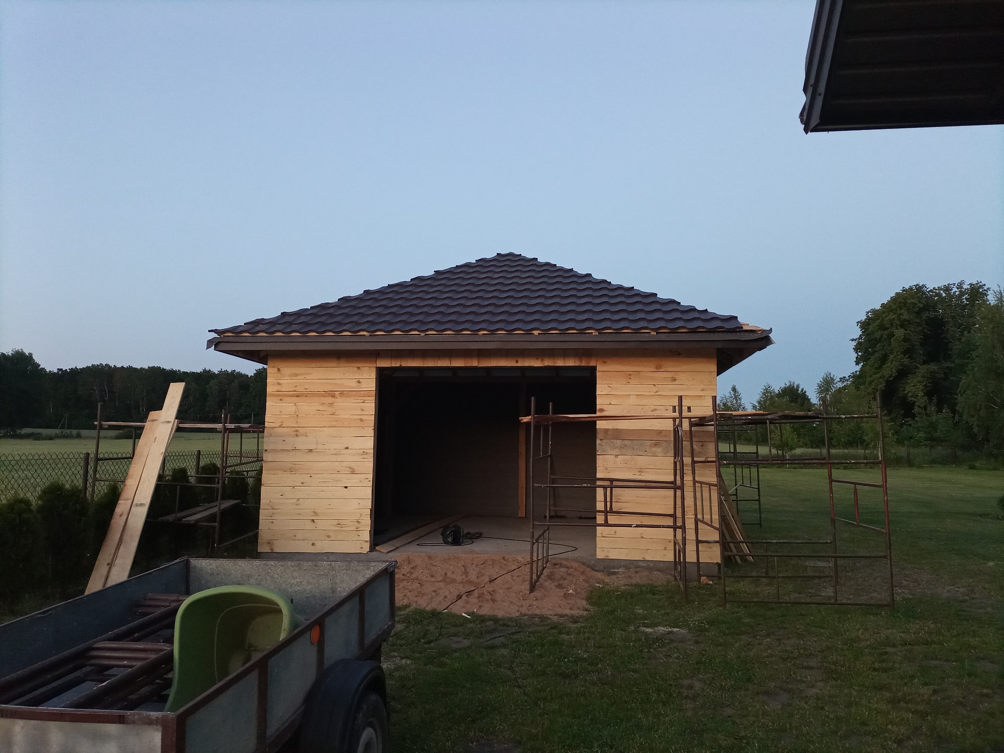 Budowa garażu z drewnianą konstrukcją, pokrytego brązową blachodachówką, widoczne rusztowania i przyczepa z narzędziami w tle, krajobraz wiejski.