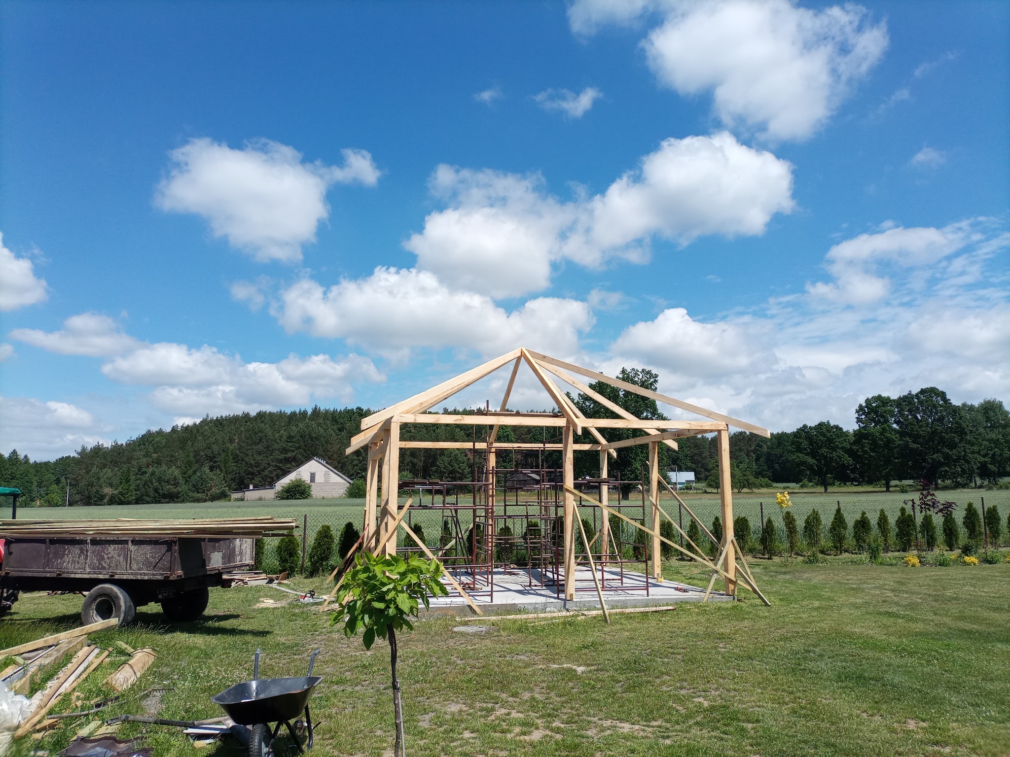 Konstrukcja drewniana w trakcie budowy na betonowej podstawie, widoczne rusztowania wewnątrz, przyczepa i taczka w tle, błękitne niebo z chmurami.