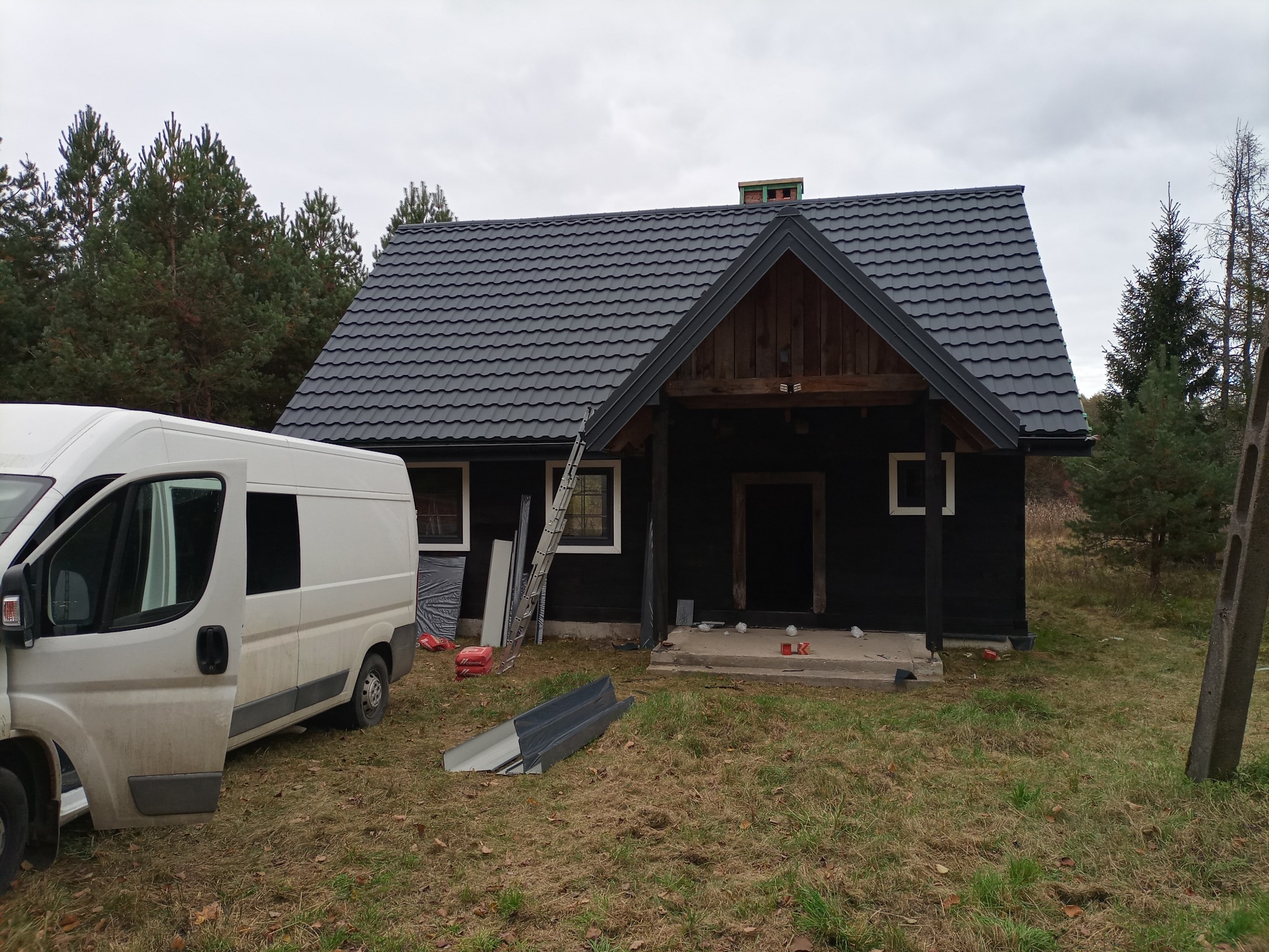 Czarny, drewniany dom z nowym, ciemnoszarym dachem z blachodachówki, drabina oparta o ścianę, biały, brudny bus z otwartymi drzwiami na trawniku, fragmenty rynny na ziemi.