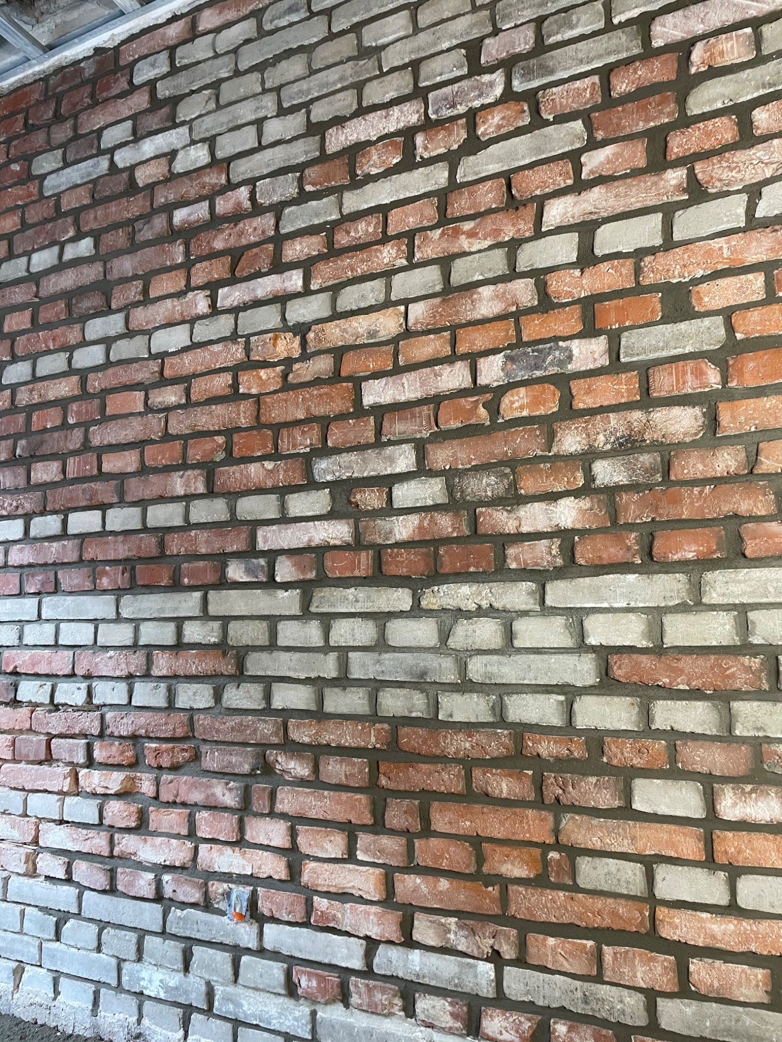 Ściana z cegły klinkierowej w odcieniach czerwieni i szarości, z wyraźnie zaznaczonymi spoinami, wykonana wewnątrz pomieszczenia, widoczne fragmenty sufitu.