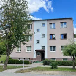 Wielorodzinny budynek mieszkalny po renowacji elewacji, z pomalowaną częścią fasady na biało i różowo, widoczny zieleń drzew i krzewów.