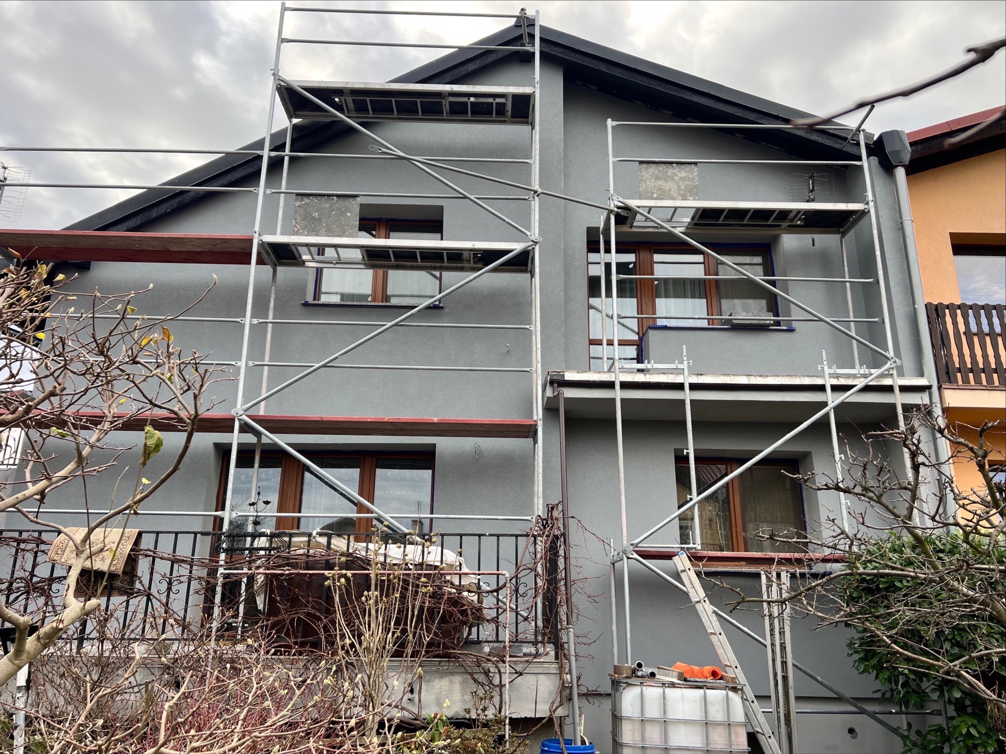 Dom w trakcie prac elewacyjnych, widoczne rusztowania, szara elewacja, brązowe ramy okienne, przygotowane powierzchnie do dalszych prac.