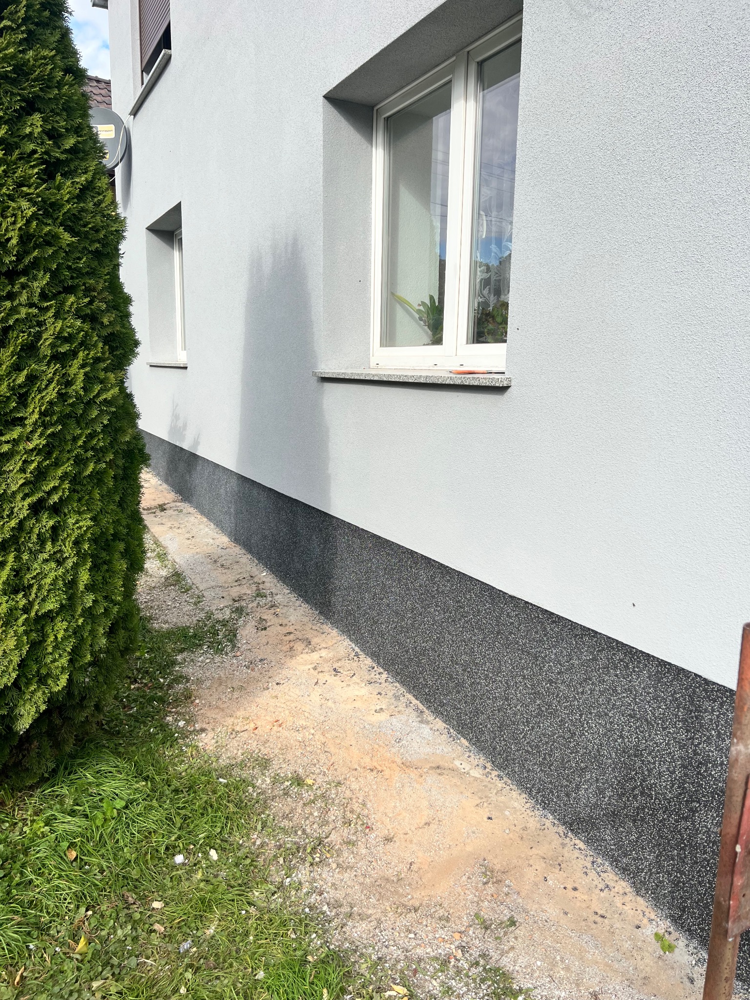 Jasnoszara ściana budynku z wykończonym cokołem w kolorze ciemnego grafitu, widoczne dwa okna, obok iglak i trawnik z piaskiem.