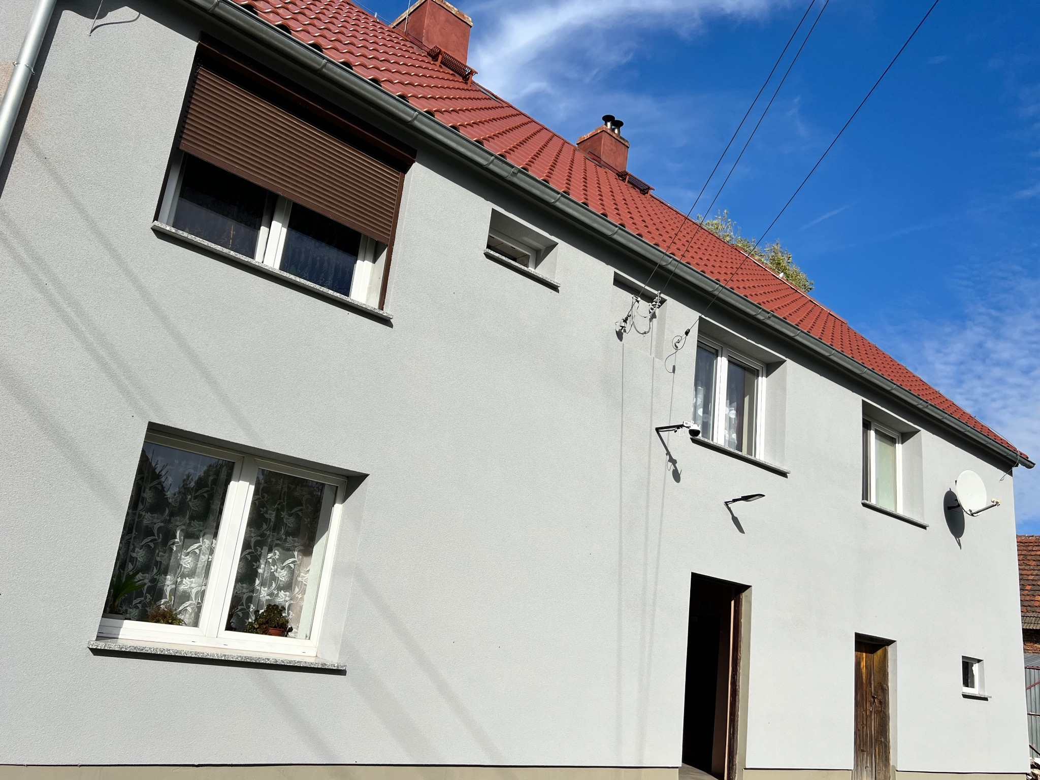 Dwukondygnacyjny budynek z szarą elewacją, czerwonym dachem i brązowymi roletami, widok z dołu na tle błękitnego nieba.