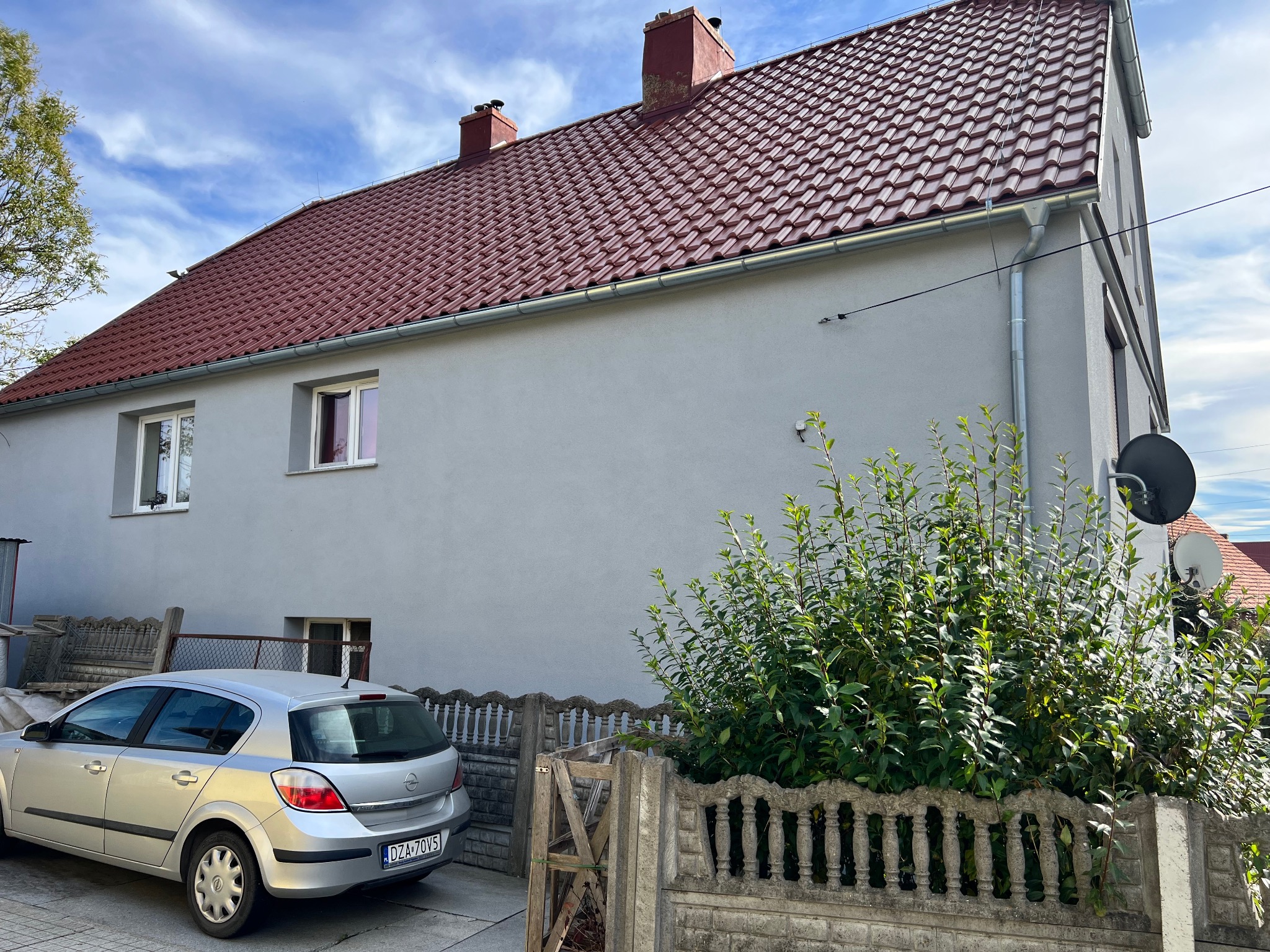 Szary dom jednorodzinny z czerwoną dachówką, widoczny srebrny samochód osobowy i betonowe ogrodzenie z zielenią.