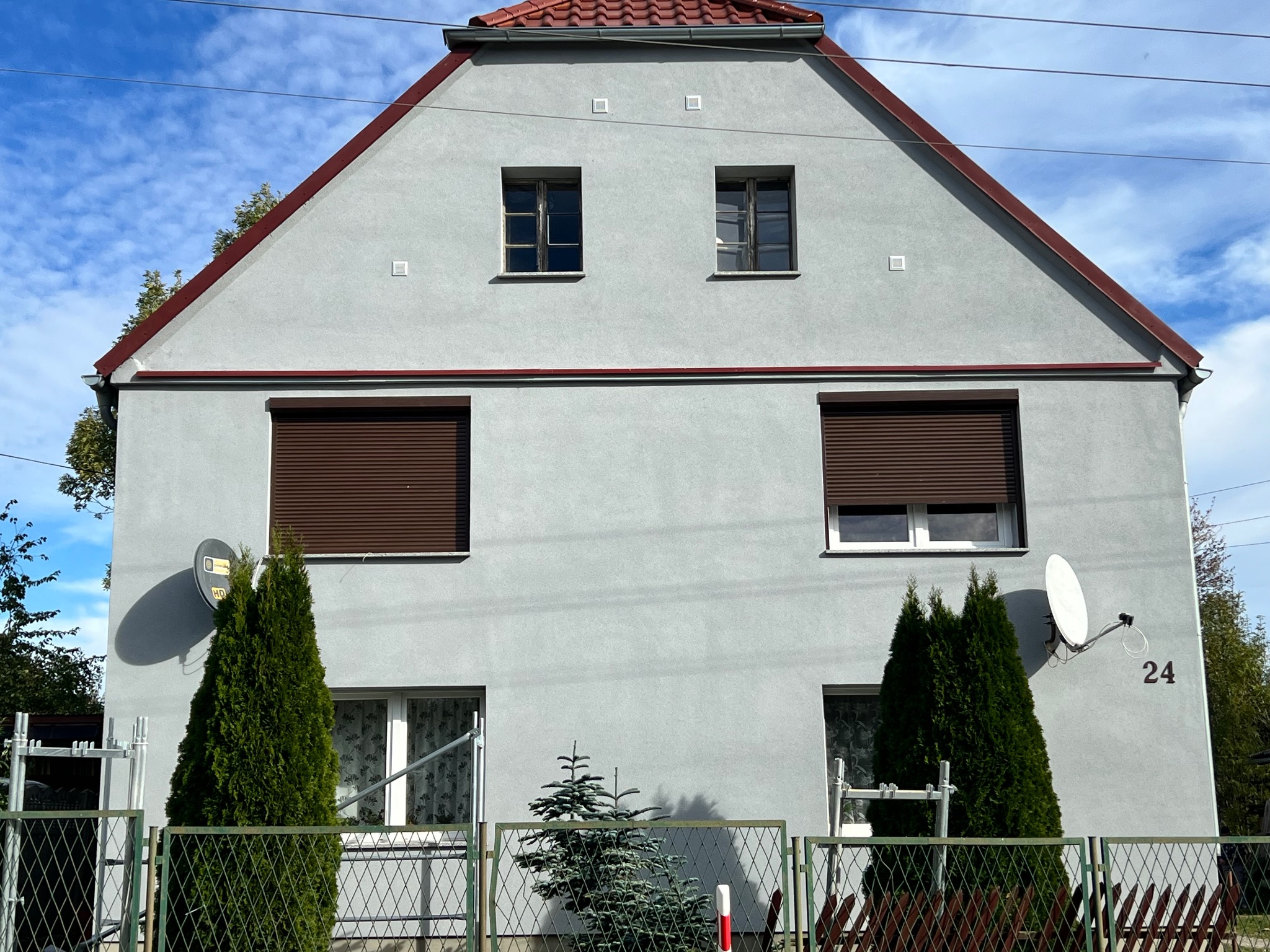 Dwupiętrowy dom z szarą elewacją, brązowymi roletami i czerwoną dachówką, widziany zza zielonego ogrodzenia z tujami i anteną satelitarną na ścianie.