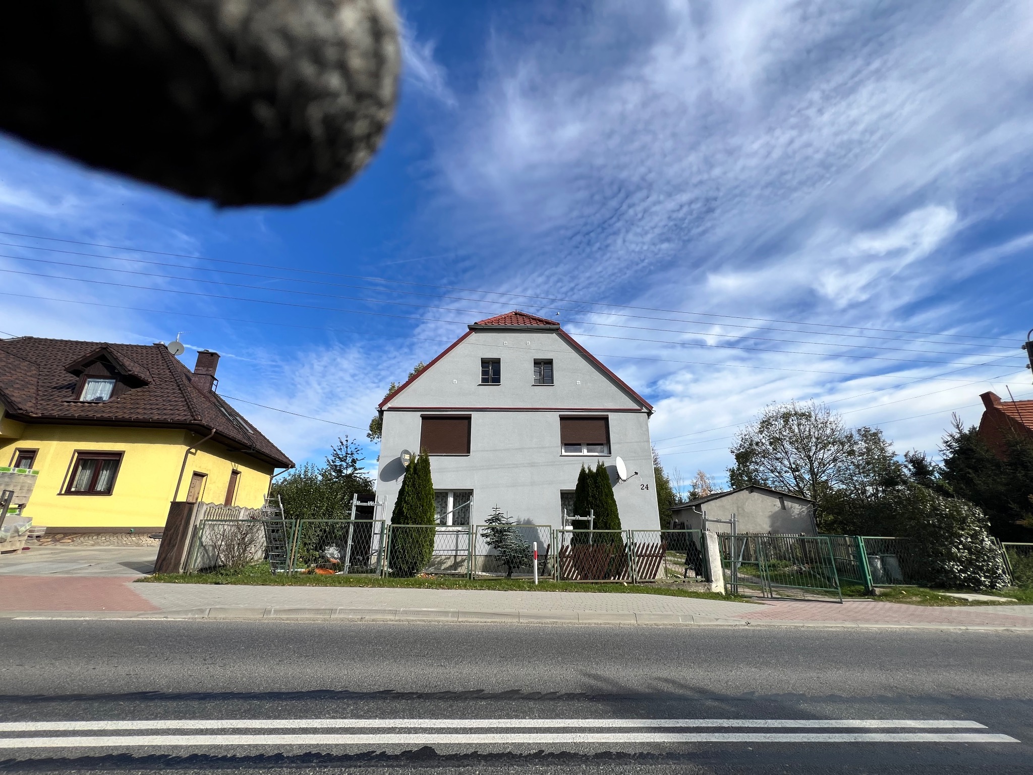 Szary dom jednorodzinny z czerwoną dachówką, widoczny od strony ulicy, z fragmentem nieostrego obiektu w górnym lewym rogu kadru.