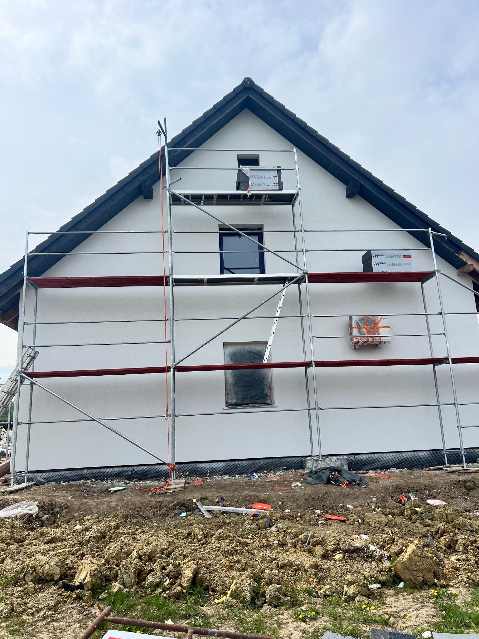 Dom jednorodzinny w trakcie prac elewacyjnych, widoczne rusztowanie, paczka materiału izolacyjnego Swissopor Lambda i zabezpieczone okna folią.
