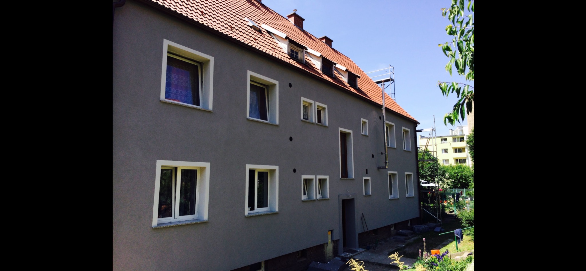 Szary budynek wielorodzinny z czerwonym dachem po remoncie elewacji, widoczne rusztowanie i fragment ogrodu w tle.