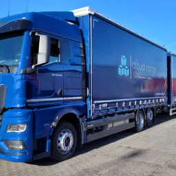Niebieski ciągnik siodłowy MAN TGX 26.480 z naczepą, oznaczony logo 'albud cargo transport & logistic', zaparkowany na brukowanym placu, widoczny fragment innego pojazdu ciężarowego.